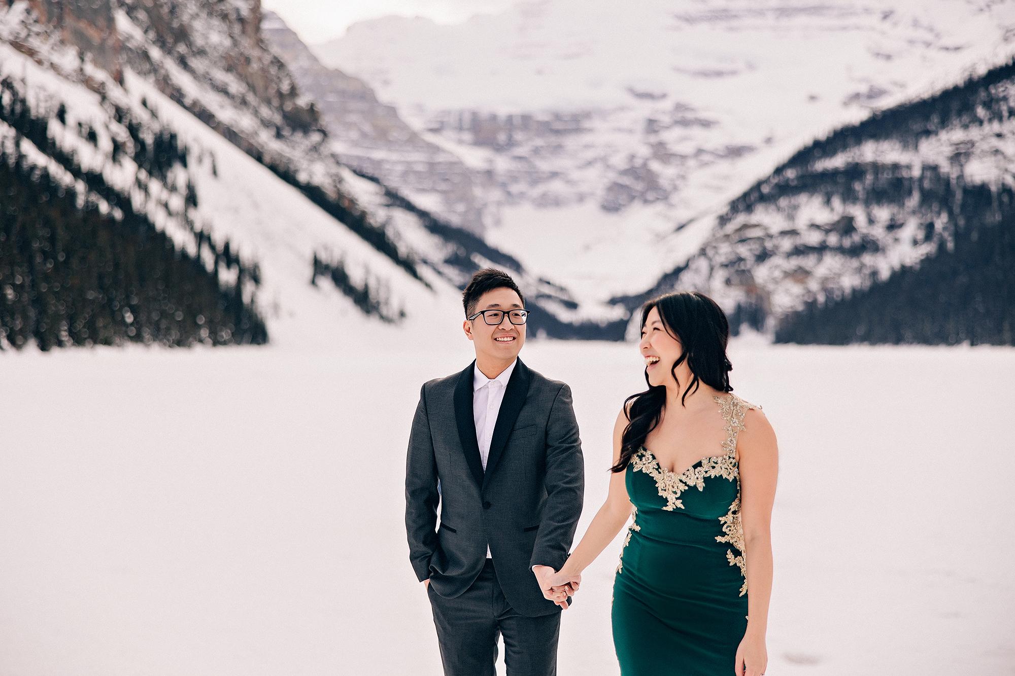 Lake Louise winter engagement photography: Joyful couple laughing and walking hand-in-hand on the frozen lake — man smiling forward, woman in emerald gold-lace gown laughing beside him — snow-covered Rocky Mountain peaks rising behind them
