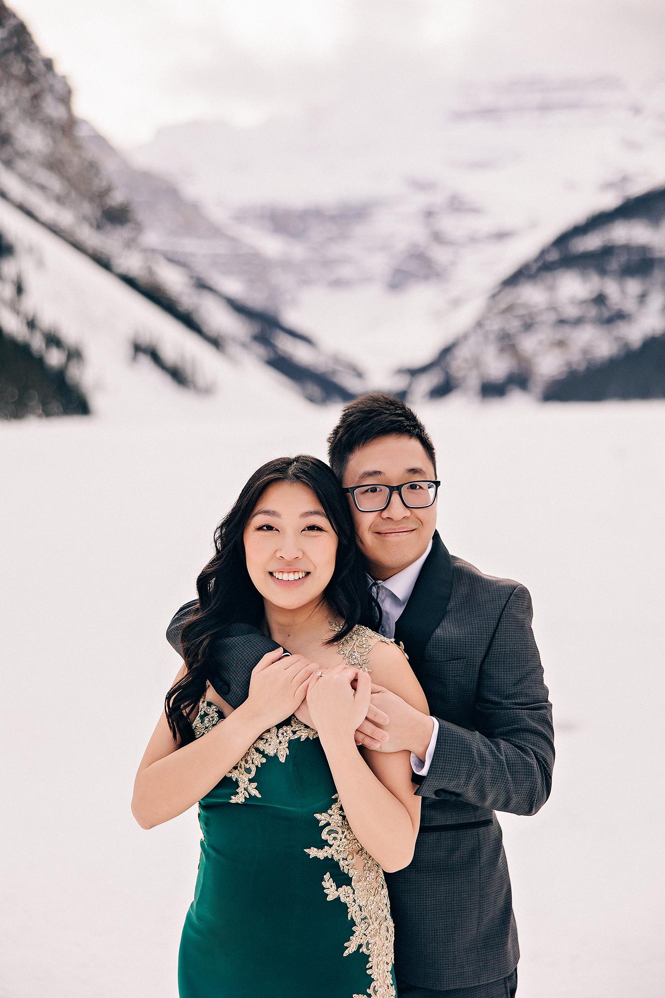 Lake Louise winter engagement photography: Tender close-up of man embracing woman from behind on the frozen lake, both smiling at camera — woman in emerald and gold-lace gown, man in dark charcoal suit — snowy Lake Louise mountain valley behind them