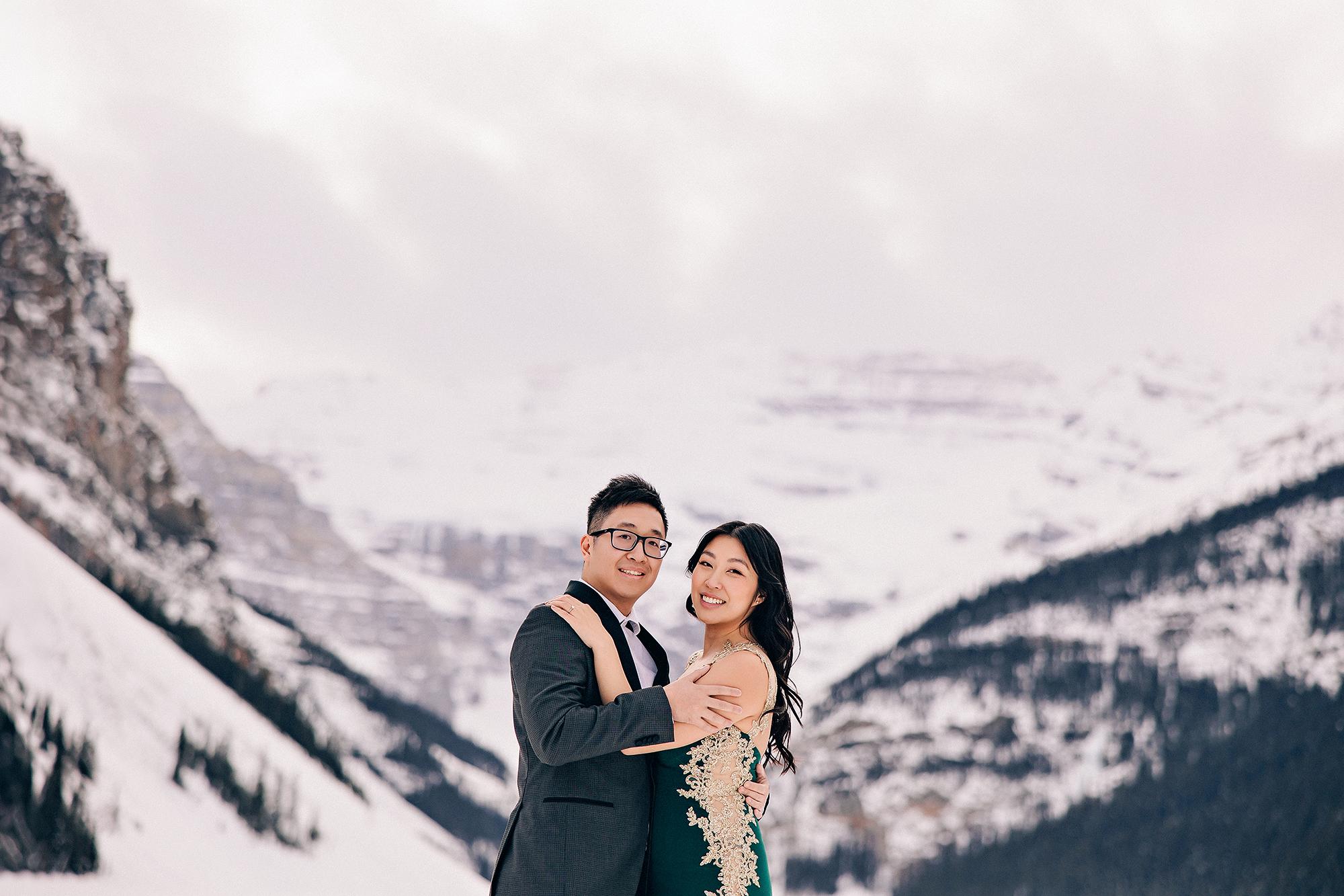 Lake Louise winter engagement photography: Couple embracing and smiling at camera on the frozen lake — woman with arm around man's shoulder in emerald gold-lace gown, man in dark charcoal suit — sweeping snow-covered Rocky Mountain peaks behind them