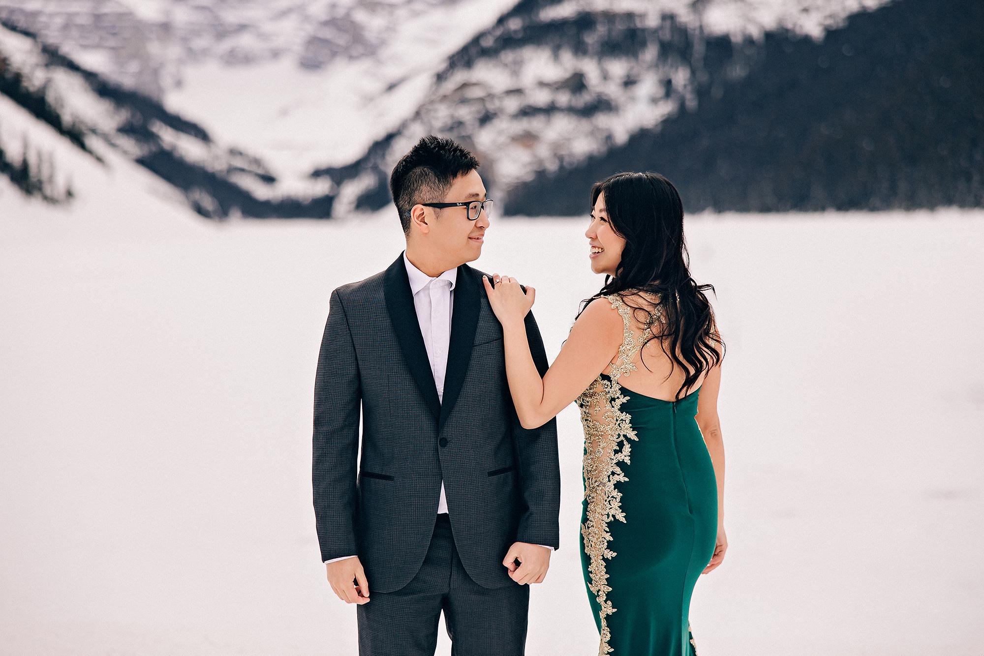 Lake Louise winter engagement photography: Intimate couple portrait on the frozen lake — woman in emerald and gold-lace backless mermaid gown touching man's shoulder, both looking at each other — snowy mountain and frozen lakeshore behind them