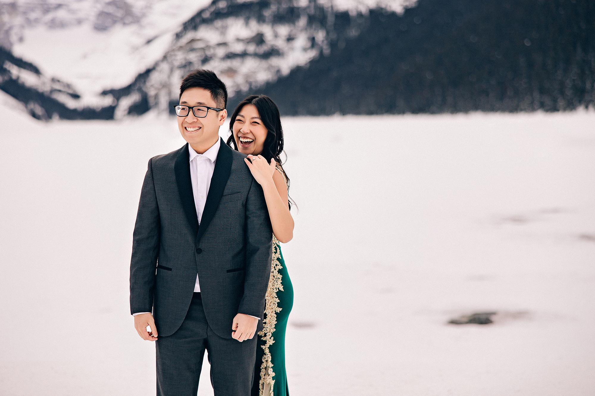 Lake Louise winter engagement photography: Joyful candid portrait of couple laughing on the frozen lake — woman in emerald gold-lace gown peeking over man's shoulder with a wide laugh, man in dark charcoal suit grinning — snow-covered Rocky Mountains behind them
