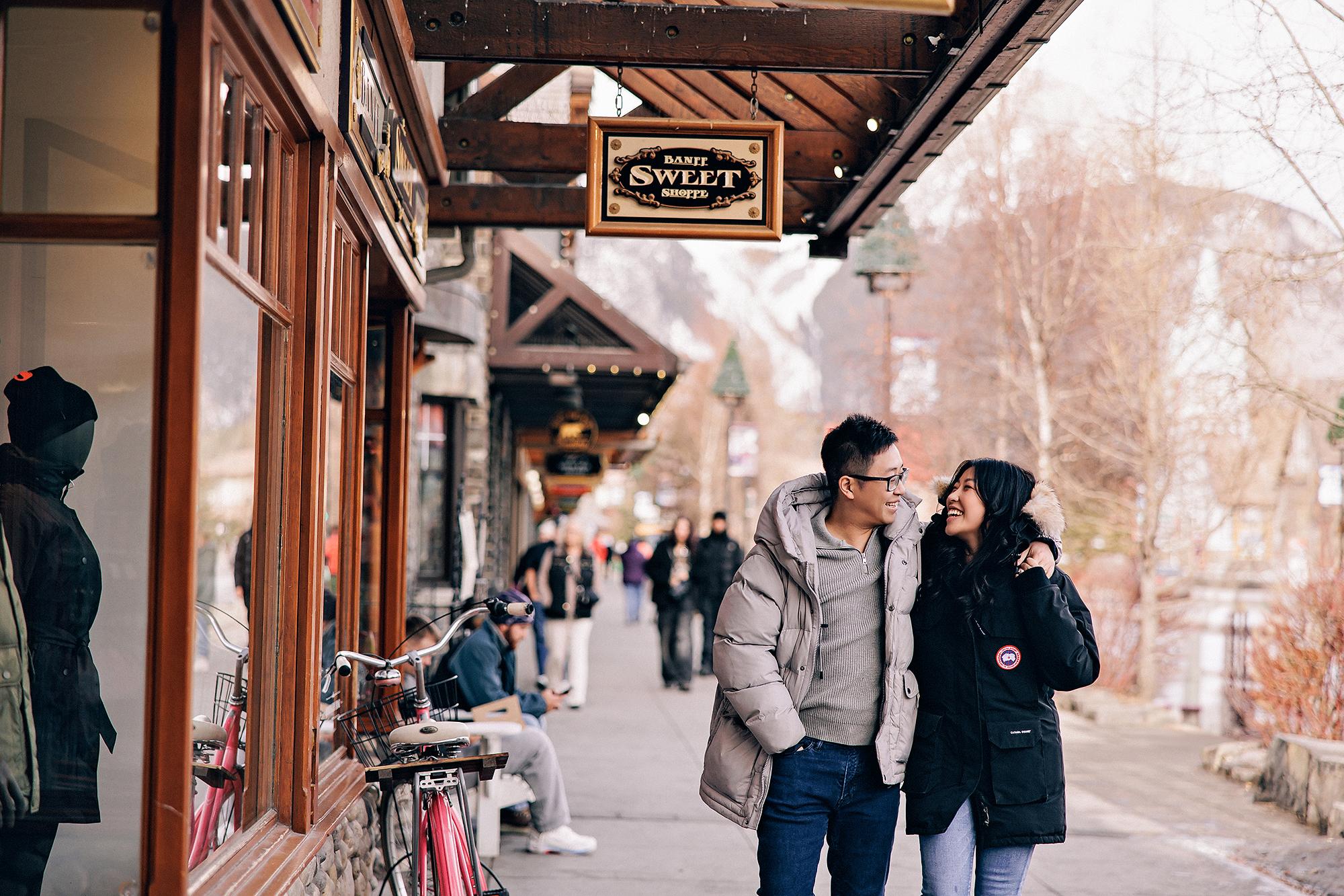 Banff engagement photography: Laughing couple in winter puffer jackets strolling along Banff Avenue under the Banff Sweet Shoppe sign, rustic storefronts and bare mountain trees behind them