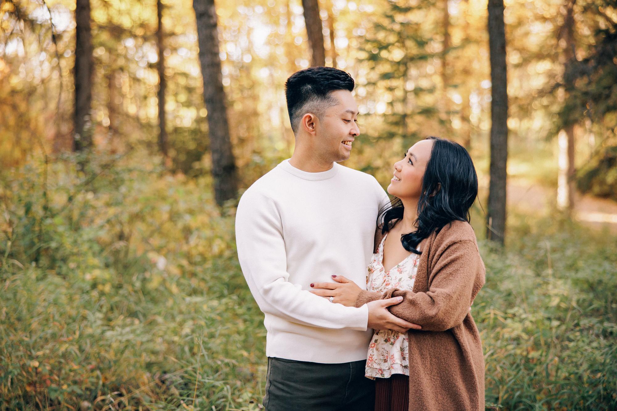 Outdoor couple photography Calgary fall: Young couple in autumn forest — man in a white crewneck sweater and woman in a floral top with a camel cardigan laughing and facing each other, golden foliage bokeh background