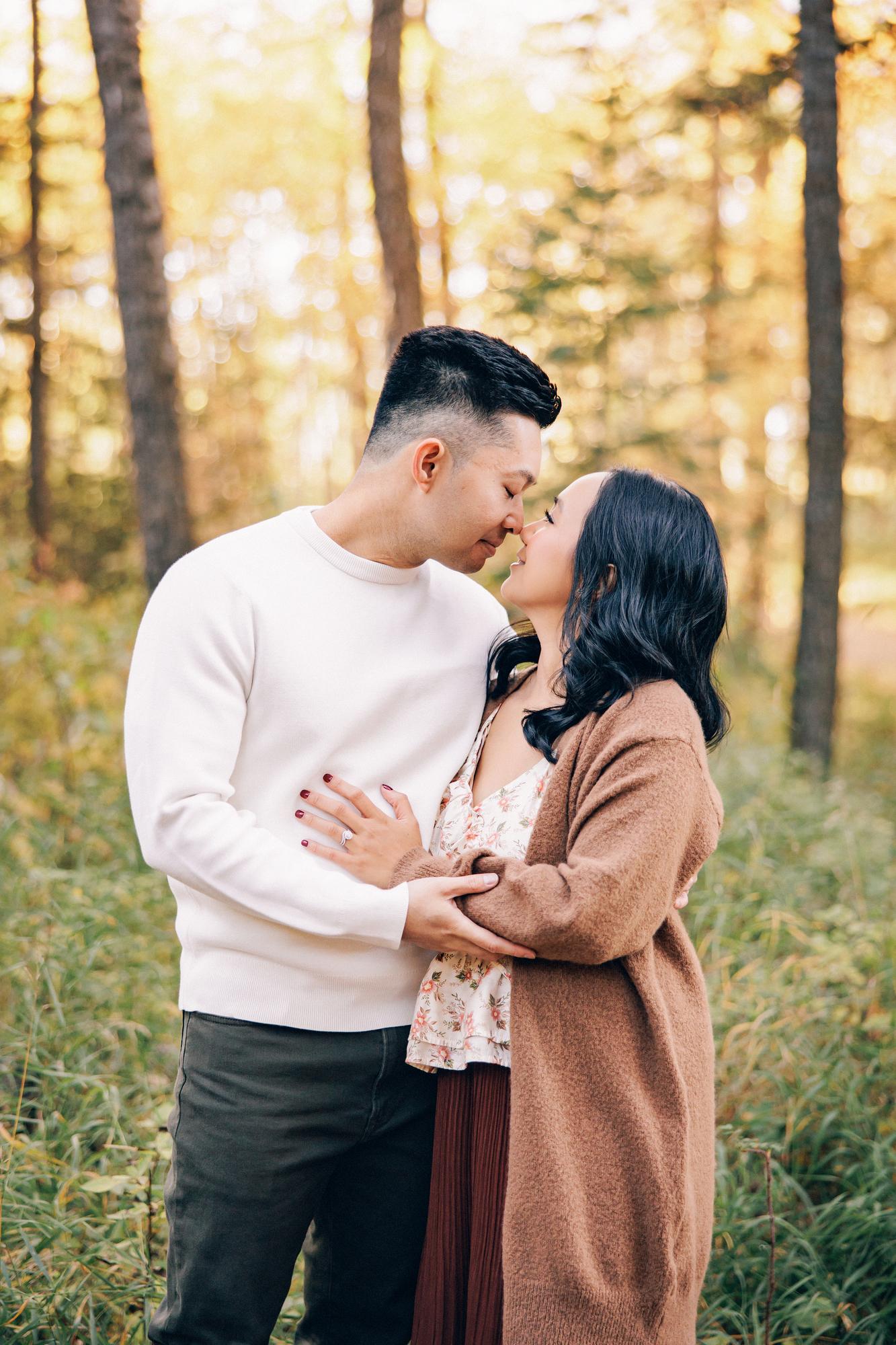 Outdoor couple photography Calgary fall: Romantic couple in autumn forest leaning in close for a near-kiss — man in white sweater, woman in floral top and camel cardigan, warm golden tree bokeh background