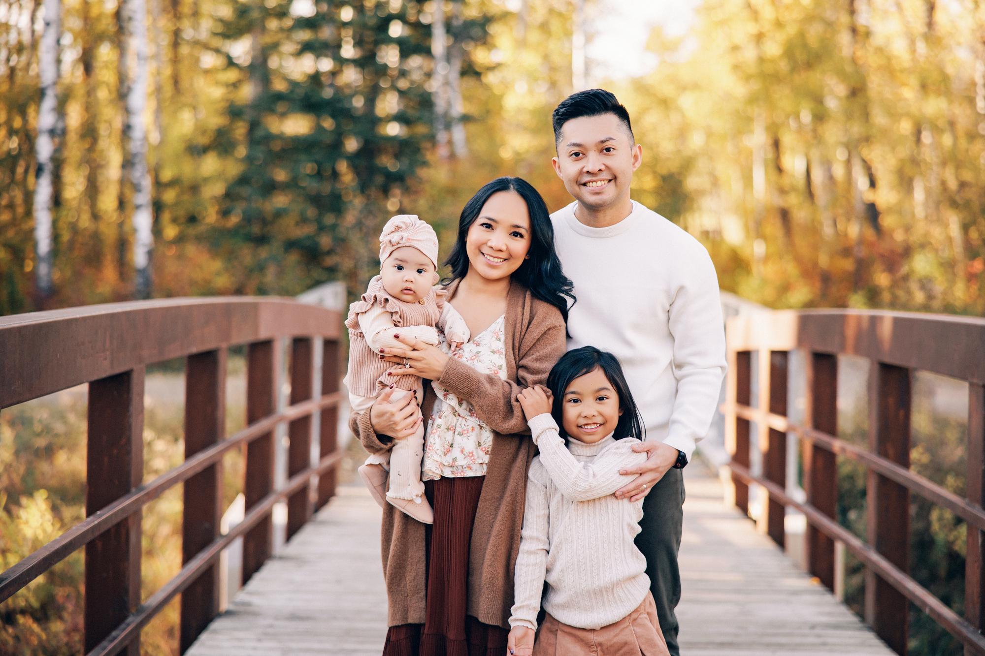 Outdoor family photography Calgary fall: Family of four on a wooden bridge — parents smiling at camera with baby girl in a pink turban and older daughter in a cream sweater, golden autumn trees behind them