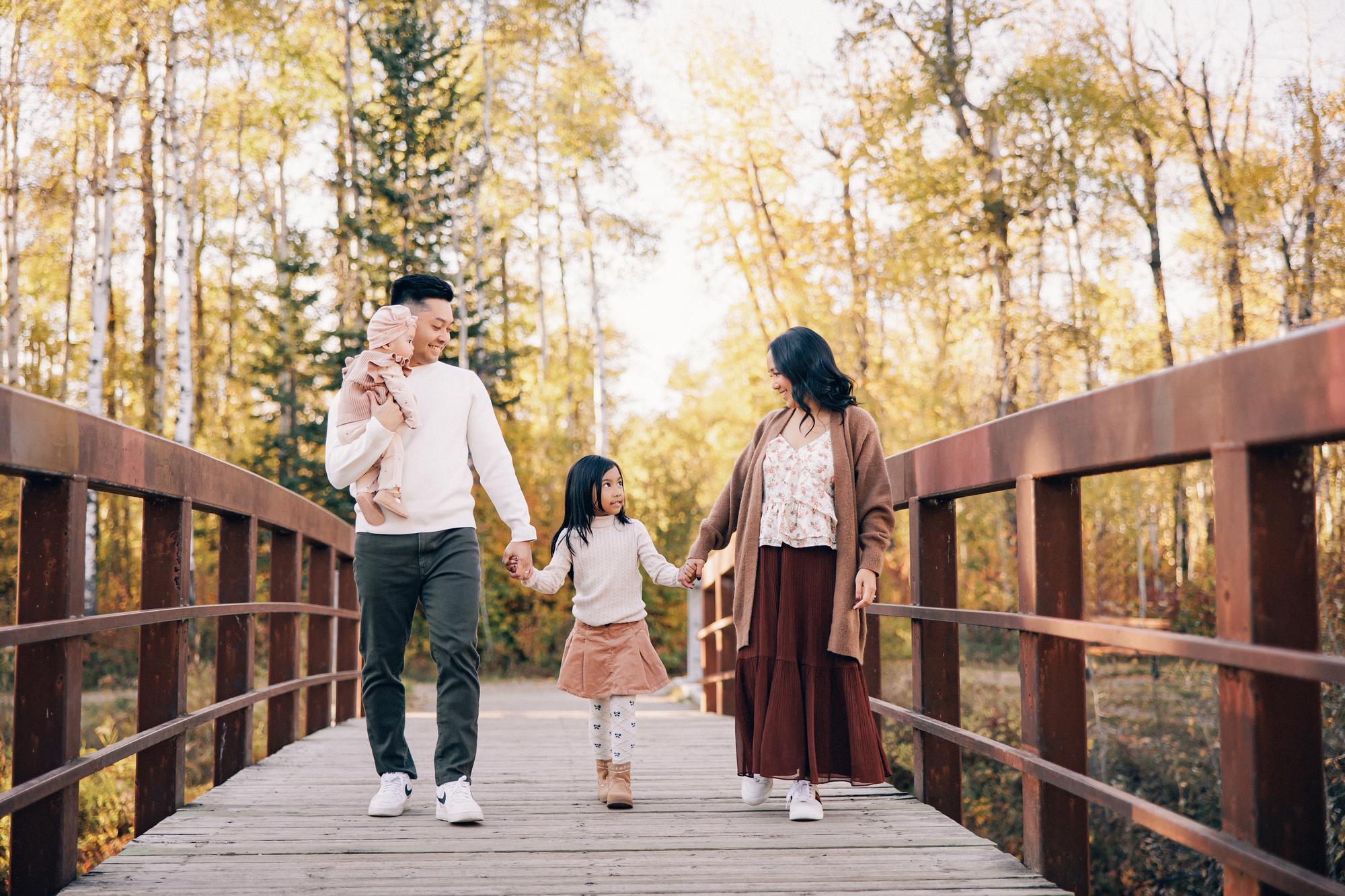 Outdoor family photography Calgary fall: Joyful family of four walking hand-in-hand across a wooden bridge — father carrying baby in pink turban, young daughter between parents, golden autumn aspens behind them