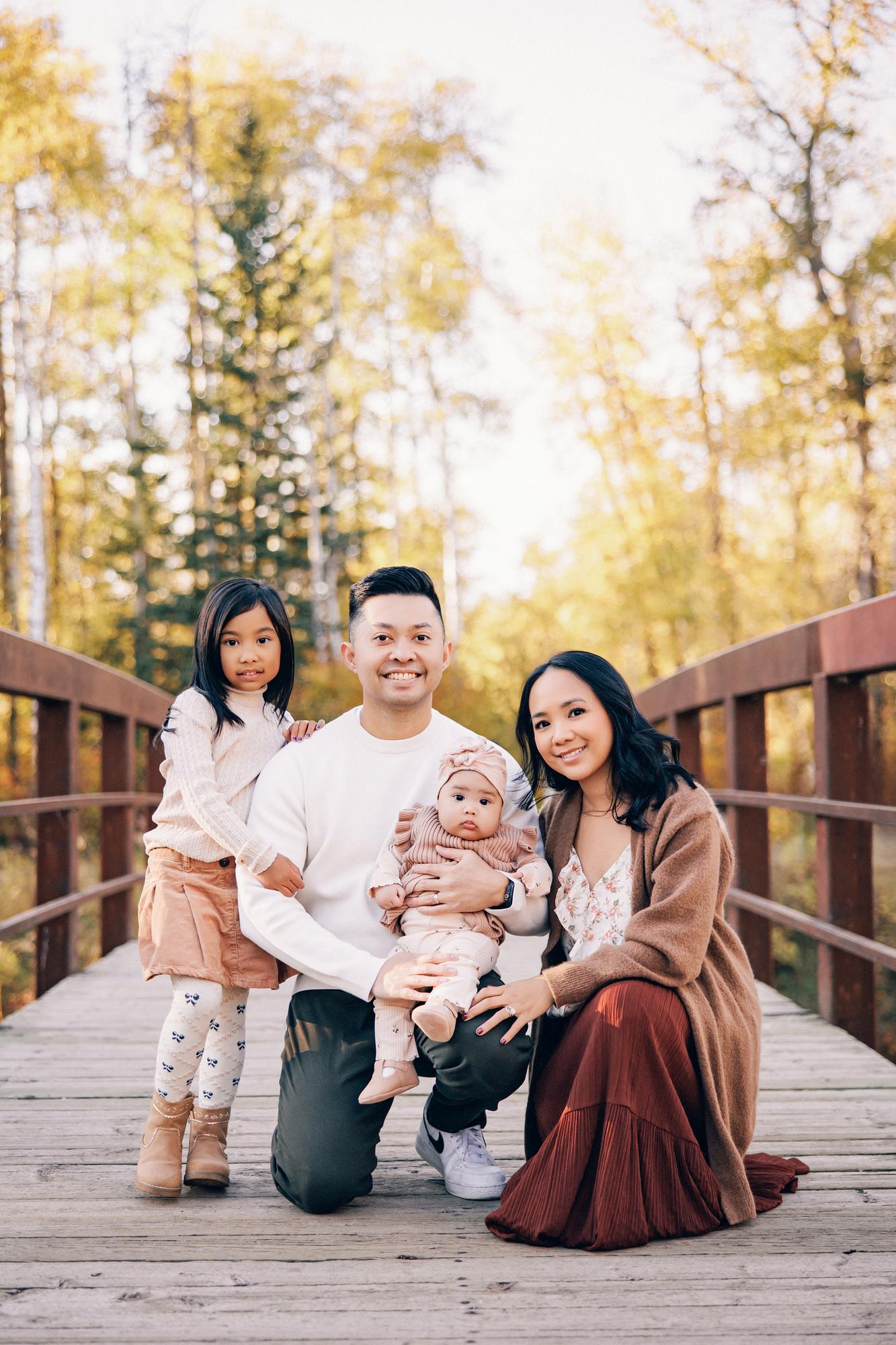Outdoor family photography Calgary fall: Family of four kneeling together on a wooden bridge — father holding baby girl in pink turban, mother beside them with young daughter standing close, surrounded by fall foliage
