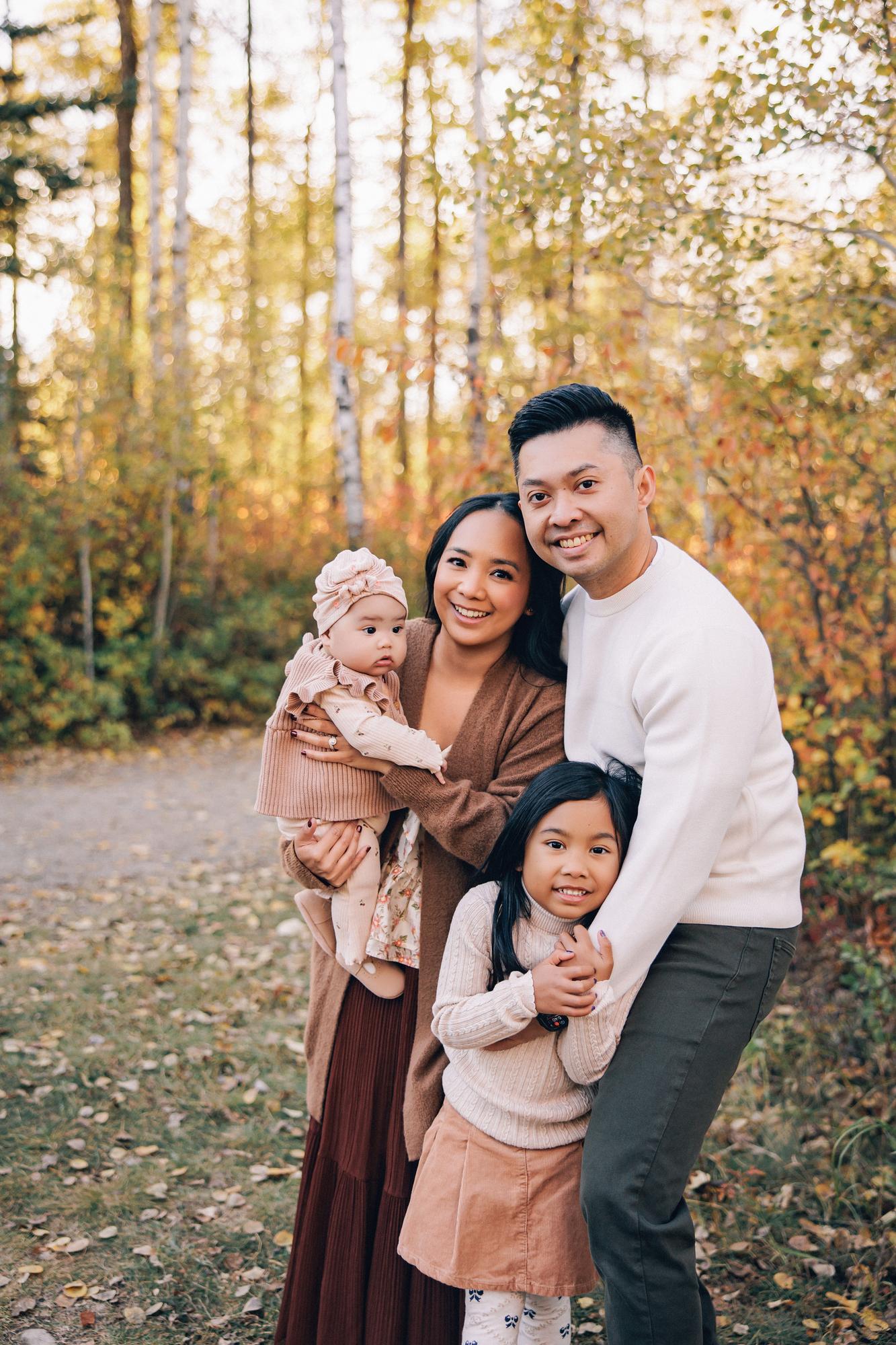 Outdoor family photography Calgary fall: Close family portrait on an autumn trail — mother holding baby girl in pink turban, father with arm around smiling young daughter, golden and rust-coloured foliage in background