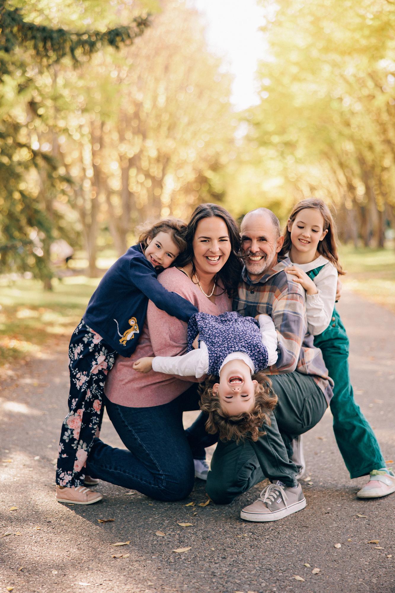 Outdoor family photography Calgary fall: Playful family of five on a tree-lined path — parents laughing while one child hangs upside-down being held, a toddler clings to mom, and an older child hugs dad, golden autumn trees behind them