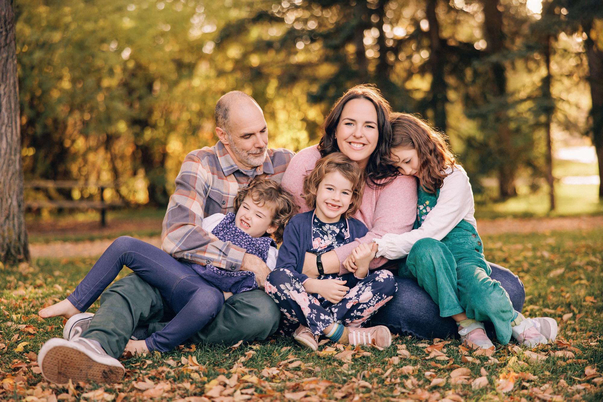 Outdoor family photography Calgary fall: Relaxed family of five seated together on grass in a sunlit park — parents and three daughters in navy, teal, and floral outfits cuddled close, golden bokeh tree background
