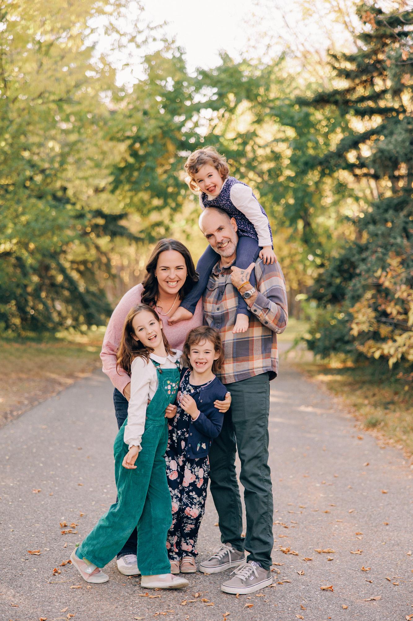 Outdoor family photography Calgary fall: Joyful family of five on a tree-lined path — mother in a pink sweater and father in a plaid flannel shirt smiling broadly, toddler on dad's shoulders, older daughter in teal corduroy overalls, and middle child in a navy floral outfit, golden autumn trees behind them