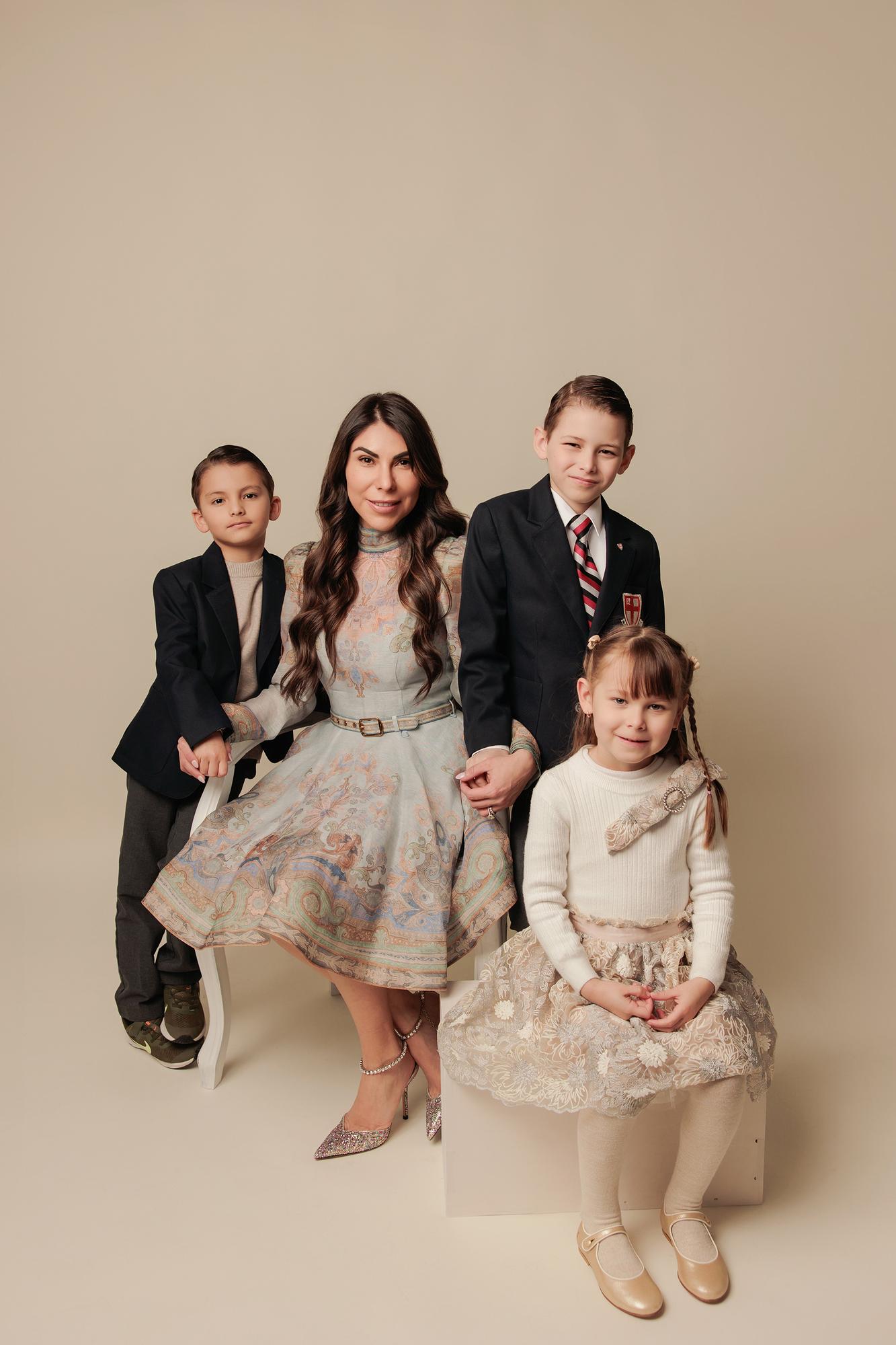 Family photography Calgary studio: Mother in a blue paisley midi dress with crystal heels seated on a white cube with three children — two boys in dark blazers and daughter in a lace skirt — warm taupe background