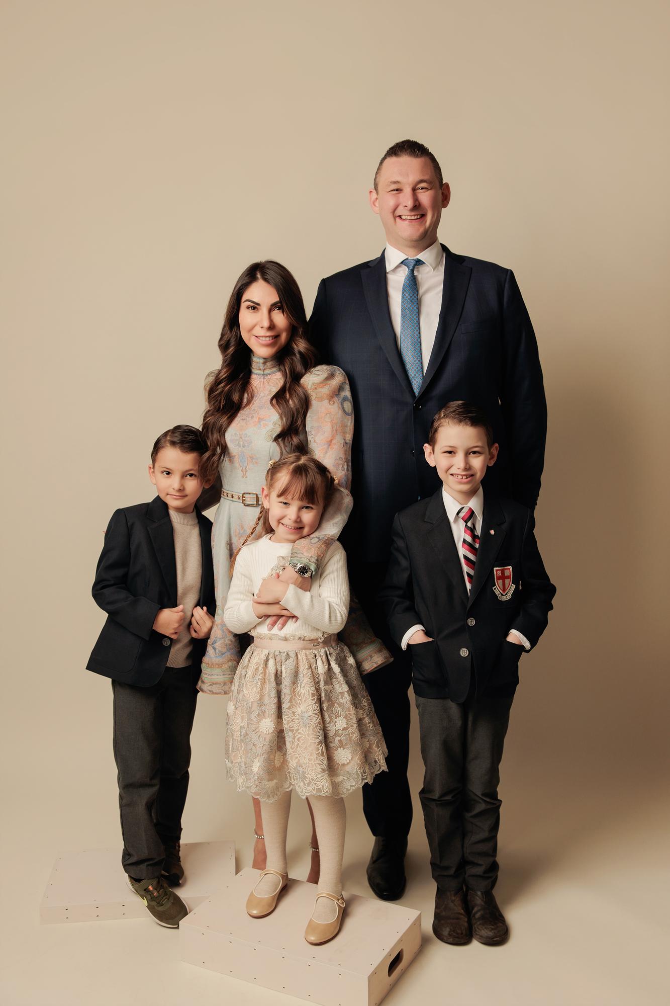 Family photography Calgary studio: Elegant family of five — mother in a blue paisley midi dress and father in a navy suit and blue tie, with two boys in dark blazers and a daughter in a lace skirt standing on a white plinth, warm taupe background