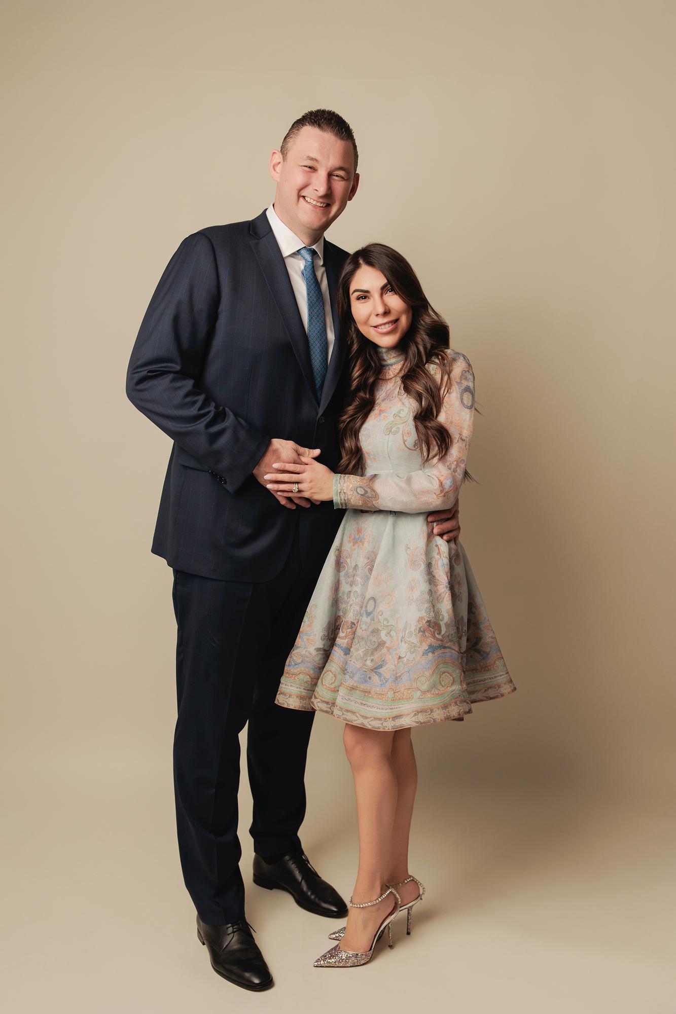 Family photography Calgary studio: Full-length couple portrait — tall man in a navy suit and blue tie with arm around woman in a blue paisley midi dress and crystal heeled sandals, warm taupe background