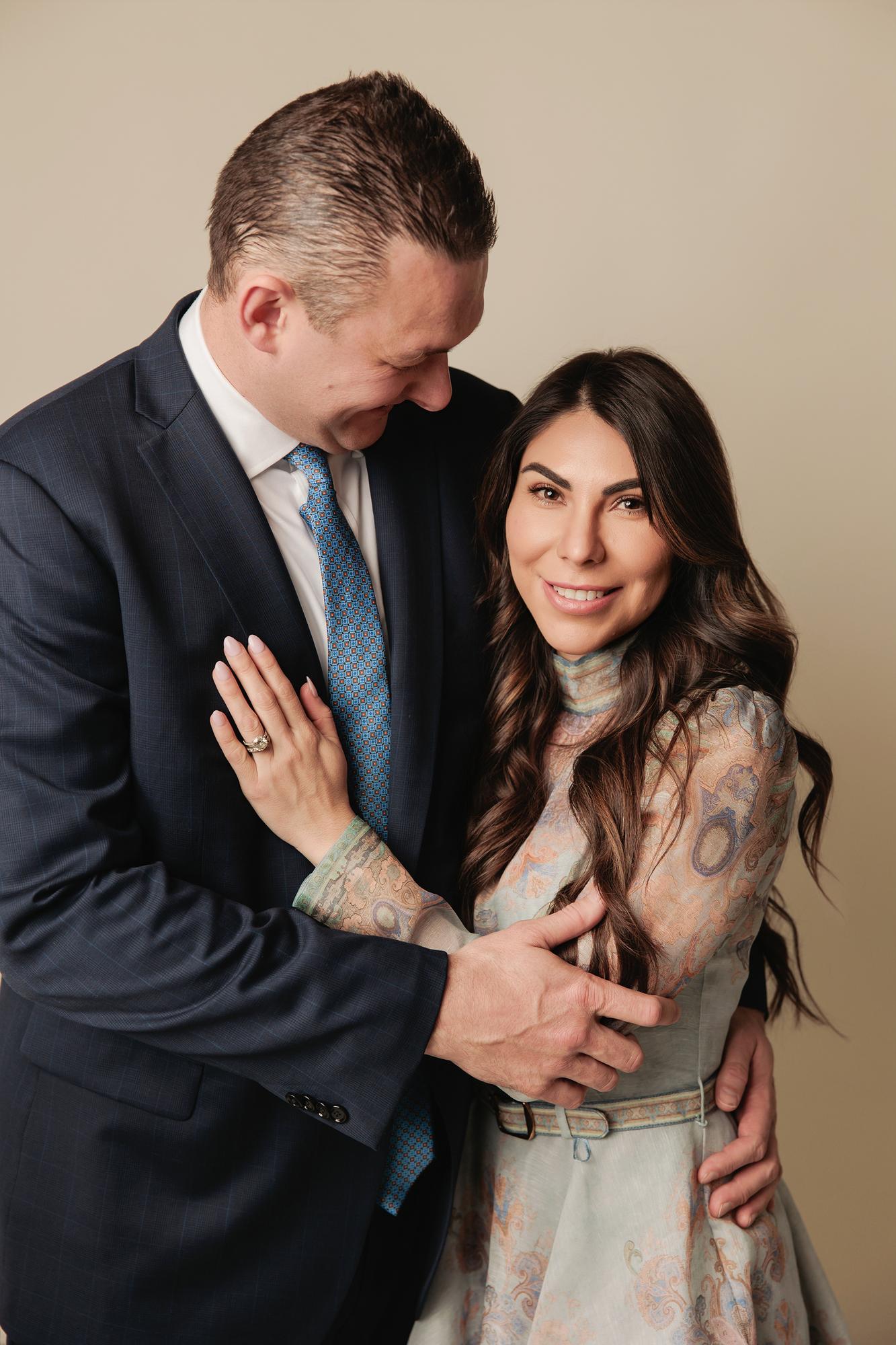 Family photography Calgary studio: Romantic close-up of couple — man in a navy suit and blue geometric tie leaning in toward smiling woman in a blue paisley dress, warm taupe background