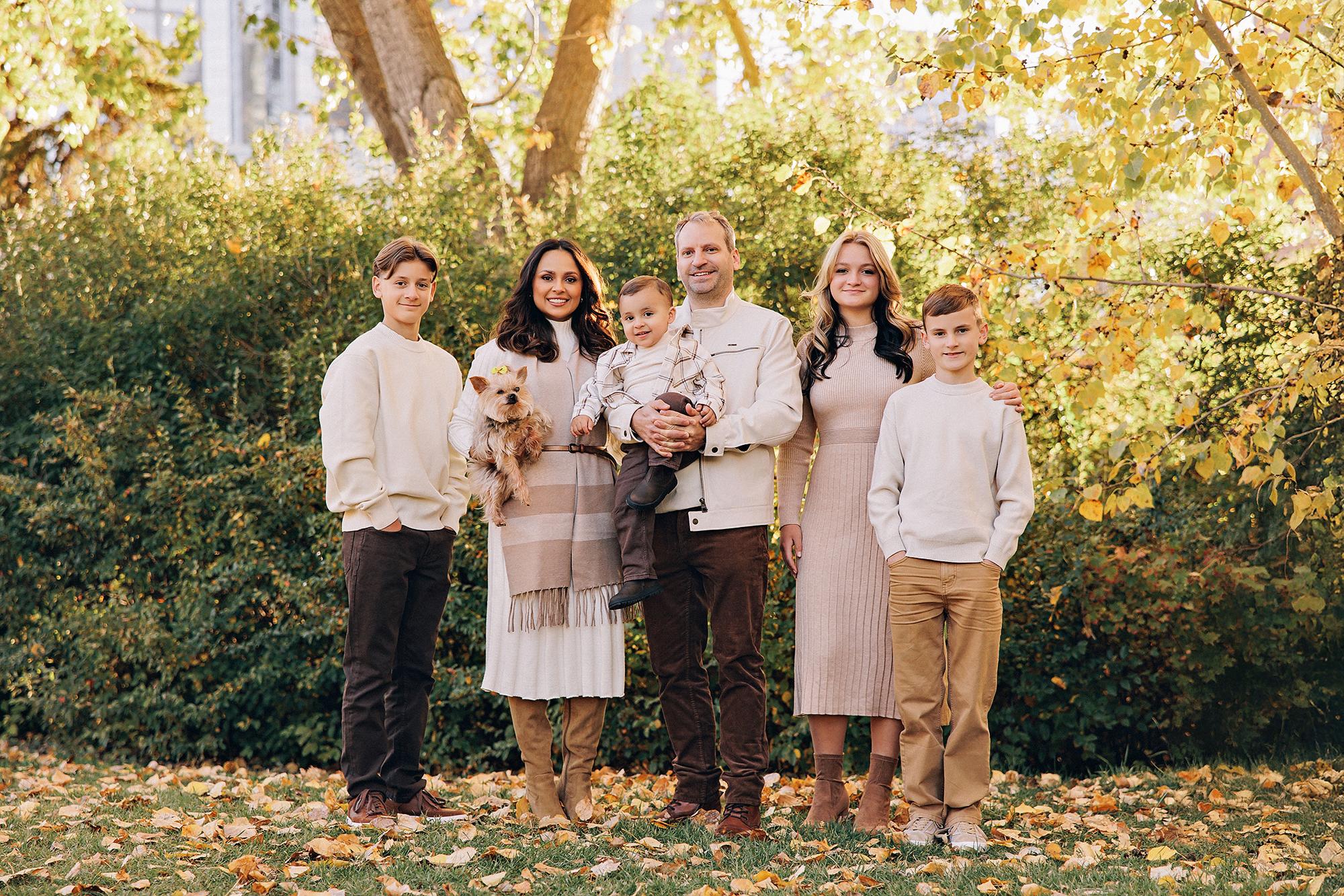 Outdoor family photography Calgary fall: Large blended family of six in coordinated cream and neutral tones — mother holding Yorkshire Terrier, father holding toddler boy in plaid shirt, teenage daughter, and two younger boys — surrounded by golden autumn foliage