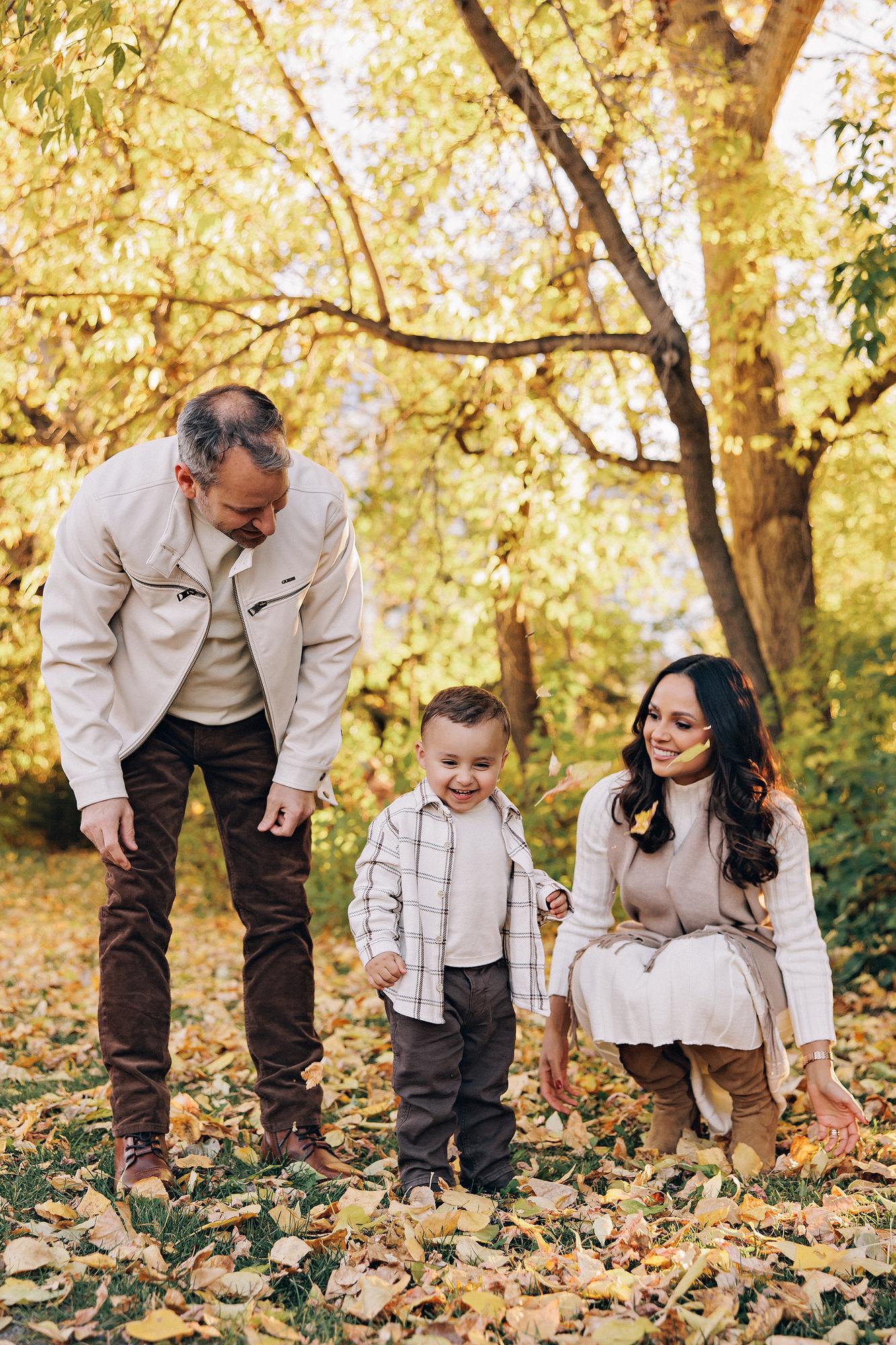 Outdoor family photography Calgary fall: Playful portrait of parents in cream outfits encouraging laughing toddler boy in plaid to walk on a path blanketed with golden autumn leaves