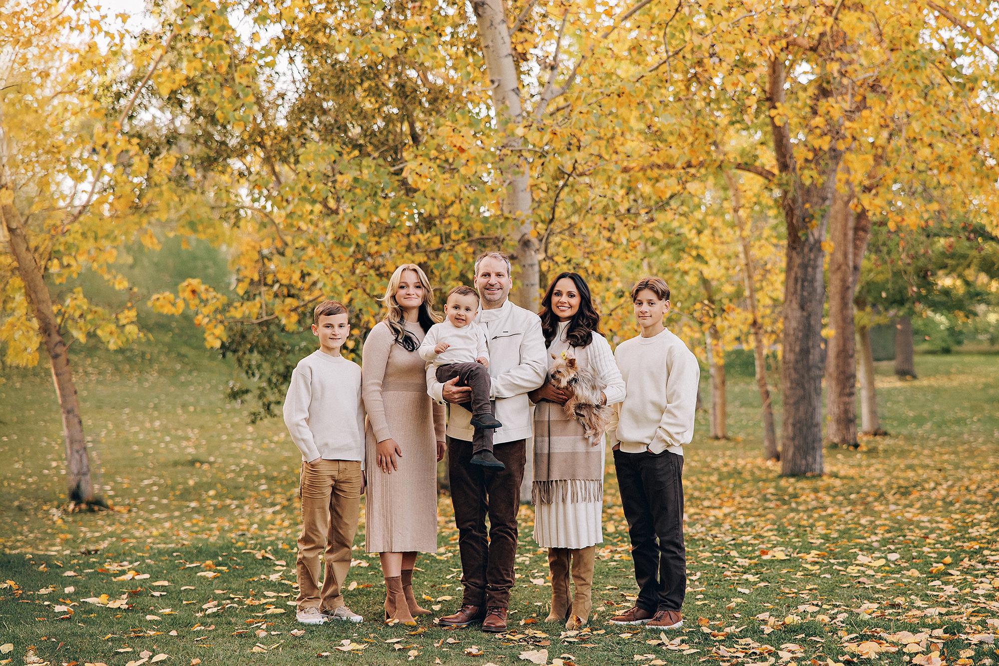 Outdoor family photography Calgary fall: Wide family portrait of six — parents in cream tones with toddler, teenage daughter, and two younger boys — standing in a golden aspen grove with fallen leaves
