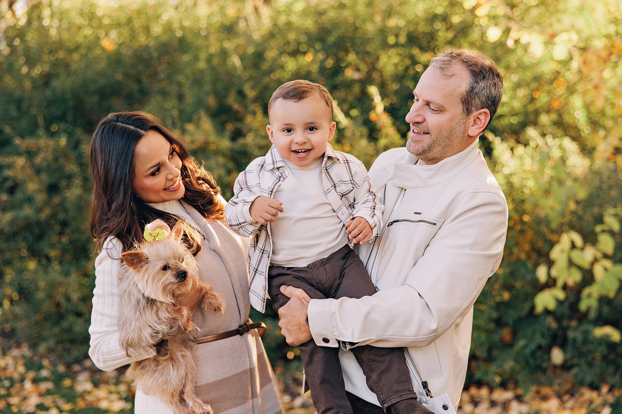 Outdoor family photography Calgary fall: Candid moment of parents in cream outfits with toddler boy in plaid laughing between them, mother holding a small Yorkshire Terrier, golden autumn leaves on the ground