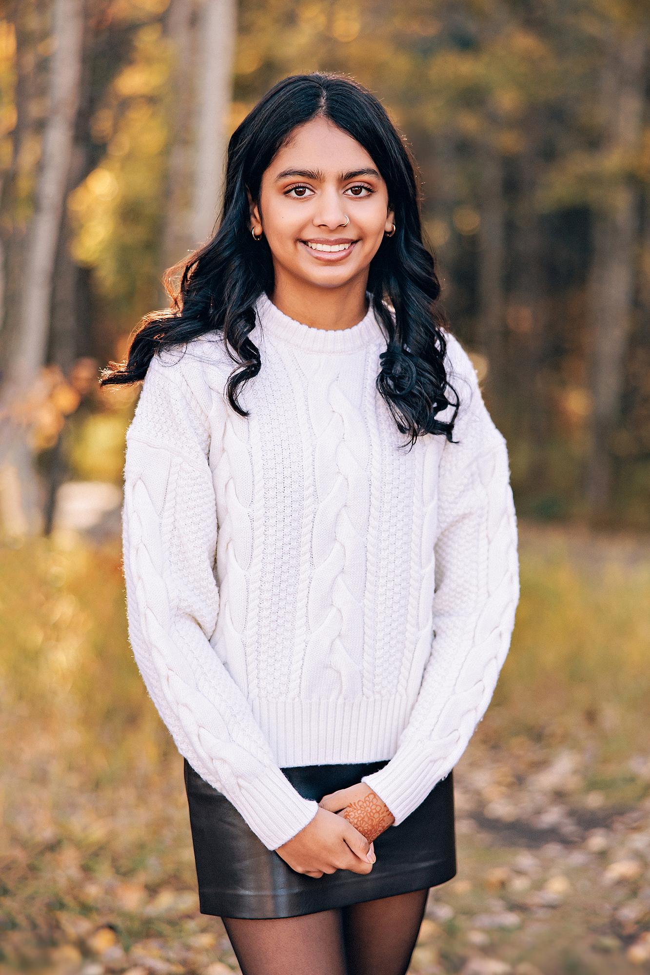 Outdoor portrait photography Calgary fall: Close-up of smiling teenage girl in a white cable-knit sweater and leather skirt with henna on her hands, golden autumn bokeh background