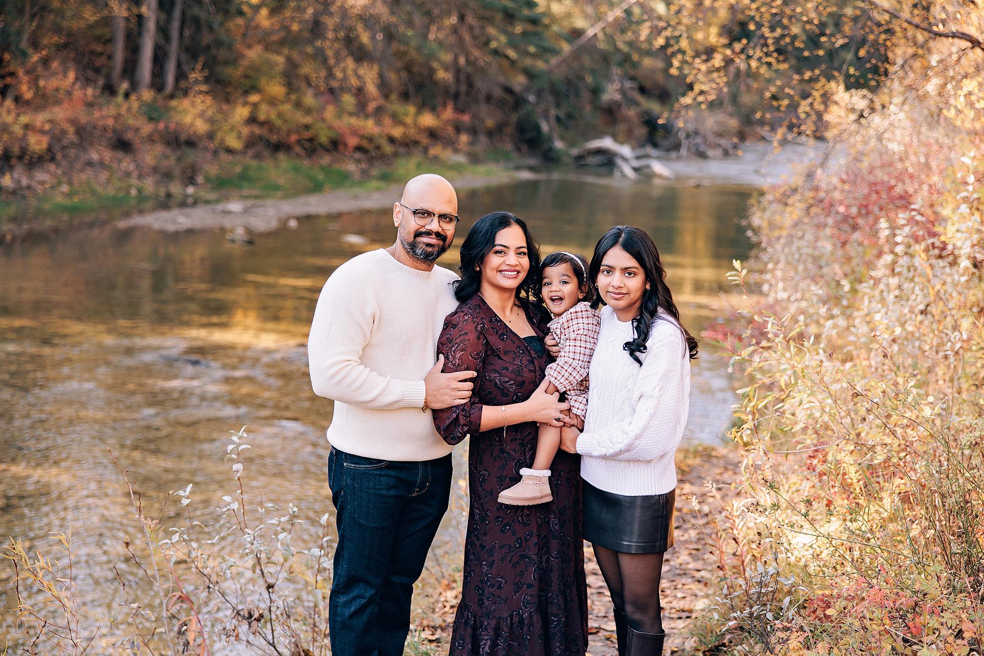 Outdoor family photography Calgary fall: Family of four by a sunlit creek — father in cream sweater and glasses, mother in burgundy floral dress holding laughing toddler in plaid, teenage daughter in white cable-knit, autumn foliage and water behind them