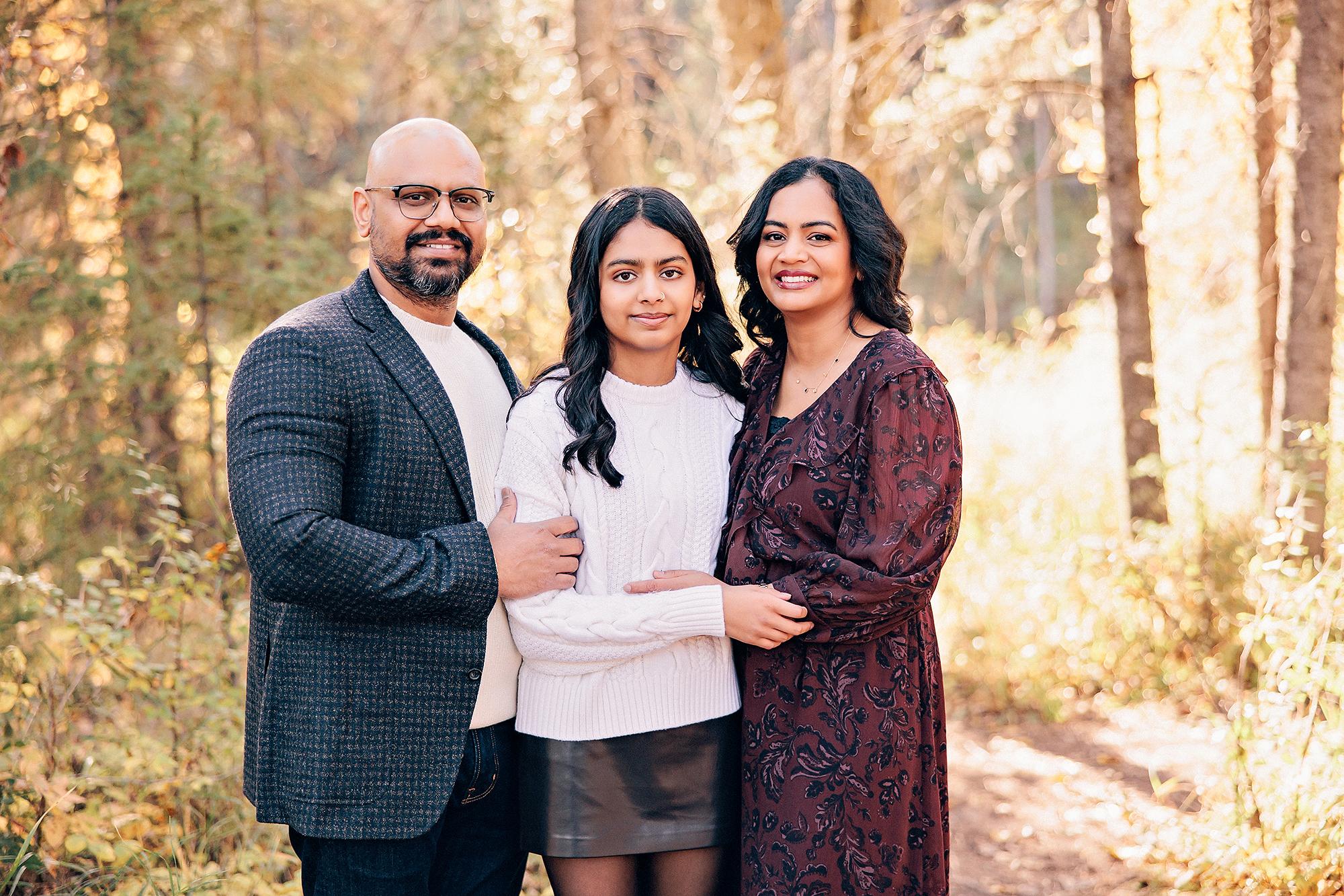 Outdoor family photography Calgary fall: Three-person portrait in a sunlit autumn forest — father in a dark houndstooth blazer, teenage daughter in white cable-knit and leather skirt, and mother in a burgundy floral dress, warm golden light