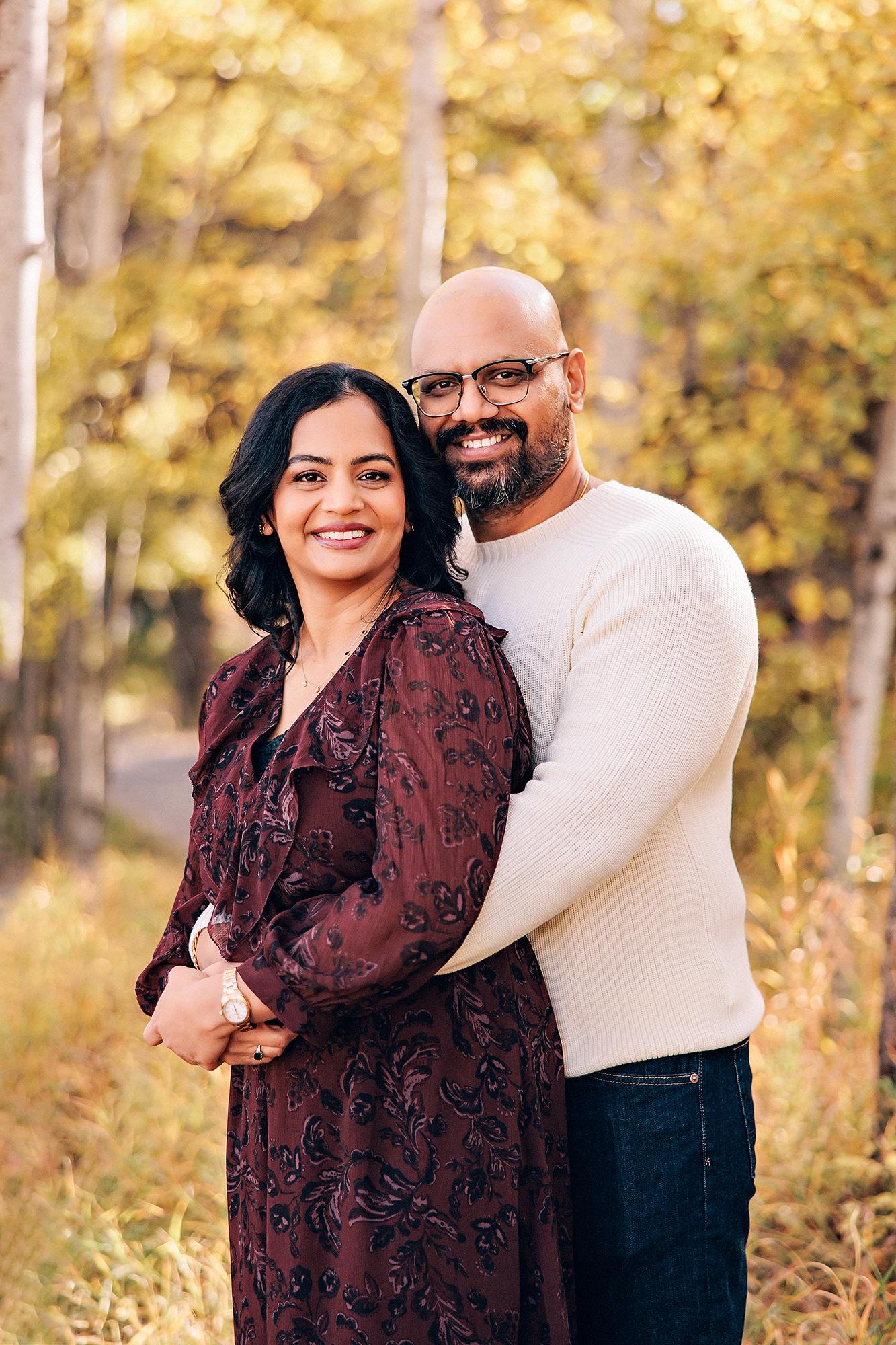 Outdoor couple photography Calgary fall: Warm portrait of couple in autumn aspen grove — woman in burgundy floral dress and man in cream ribbed sweater with glasses embracing, golden birch trees behind them