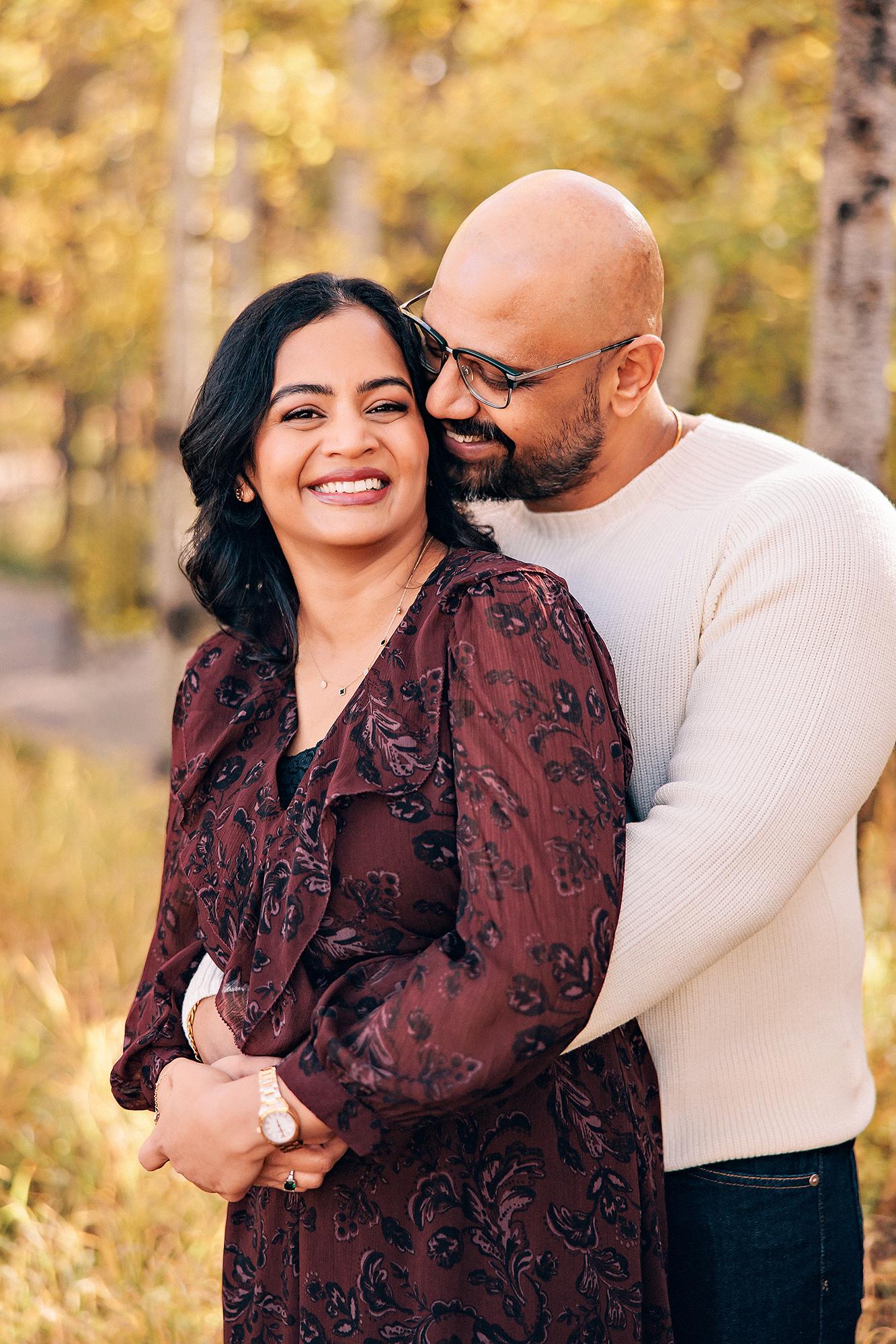 Outdoor couple photography Calgary fall: Romantic close-up of couple in autumn aspen grove — man with glasses leaning into smiling woman in a burgundy floral dress, golden bokeh foliage background