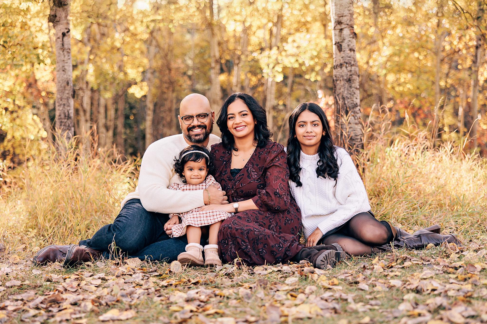 Outdoor family photography Calgary fall: Family of four seated together in golden autumn grass — father in cream sweater with toddler in plaid on his lap, mother in burgundy floral dress, and teenage daughter in white cable-knit, birch trees behind them