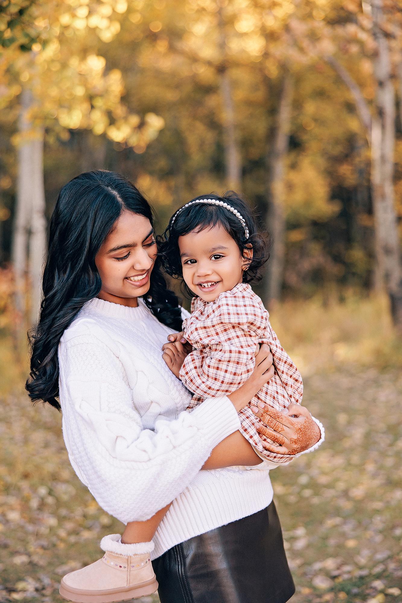 Outdoor family photography Calgary fall: Teenage daughter in a white cable-knit sweater and leather skirt holding smiling toddler sister in a plaid dress with a pearl headband, golden autumn aspen trees behind them, henna visible on her hands