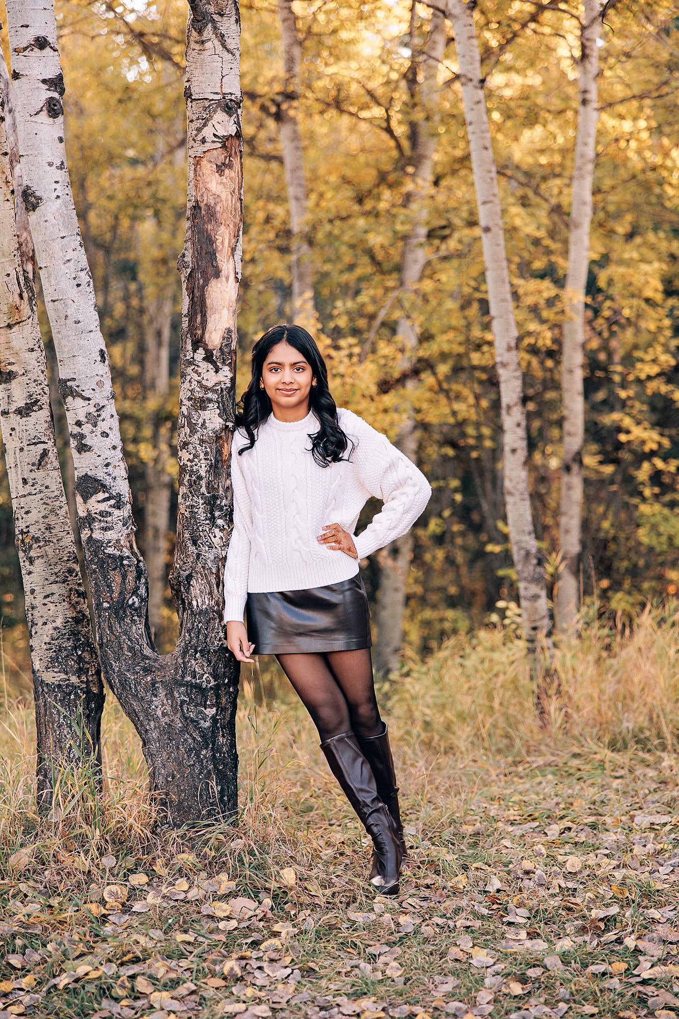 Outdoor portrait photography Calgary fall: Confident teenage girl in a white cable-knit sweater, black leather skirt, and tall black riding boots leaning against a birch tree in a golden autumn aspen grove