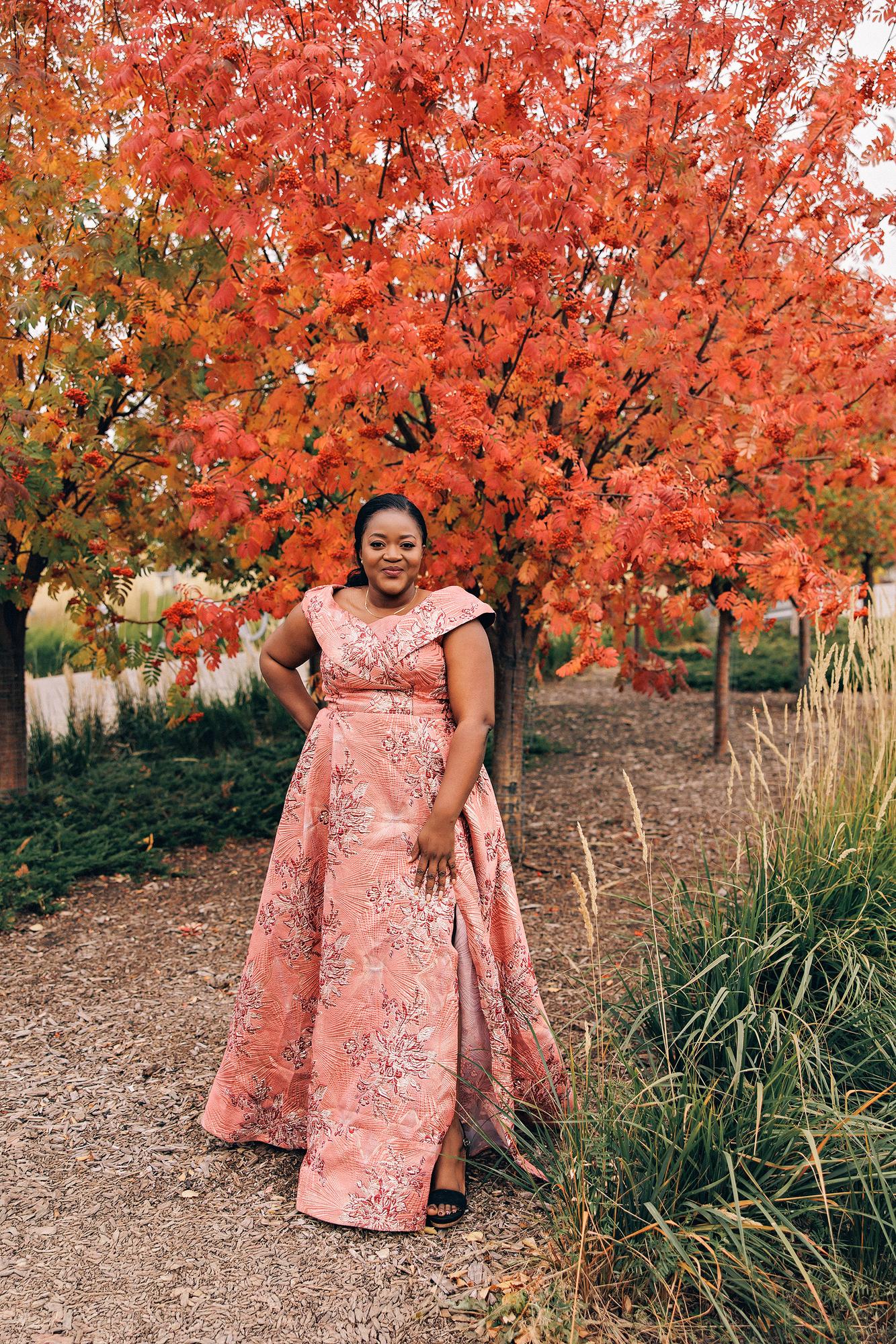 Outdoor birthday photography Calgary fall: Confident woman in a blush-pink brocade jacquard ball gown with hand on hip standing on a garden path under a vibrant red autumn mountain ash tree in full berry