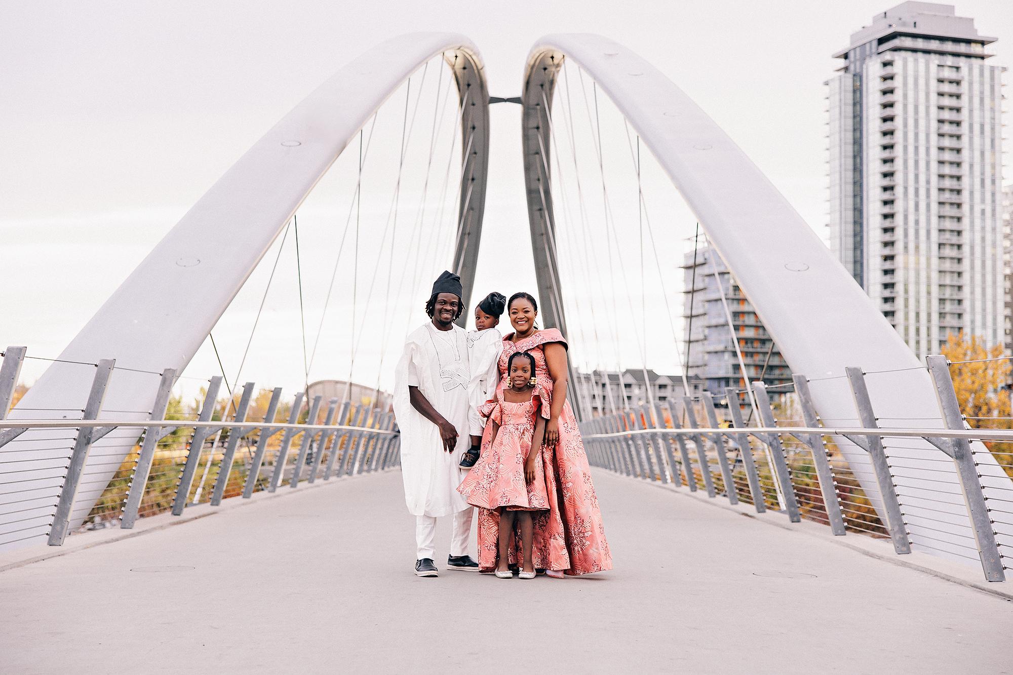 Outdoor birthday family photography Calgary: Family of four in Nigerian traditional attire — man in white agbada and fila, woman and daughter in matching pink-coral brocade gowns, and toddler boy in white — posed on a pedestrian bridge with Calgary skyline and arched cable supports behind them