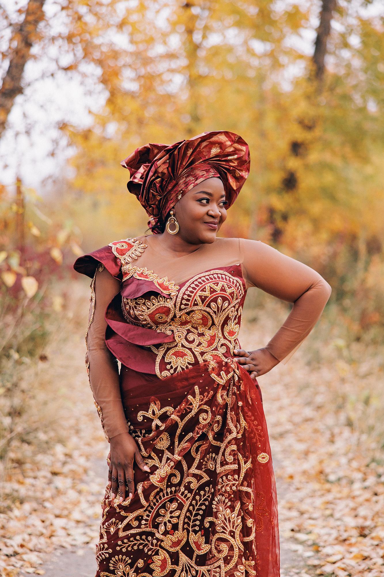Outdoor birthday photography Calgary fall: Confident woman in a burgundy and gold embroidered Nigerian lace gown with matching gele headwrap and statement gold earrings, standing with hand on hip along a golden autumn trail