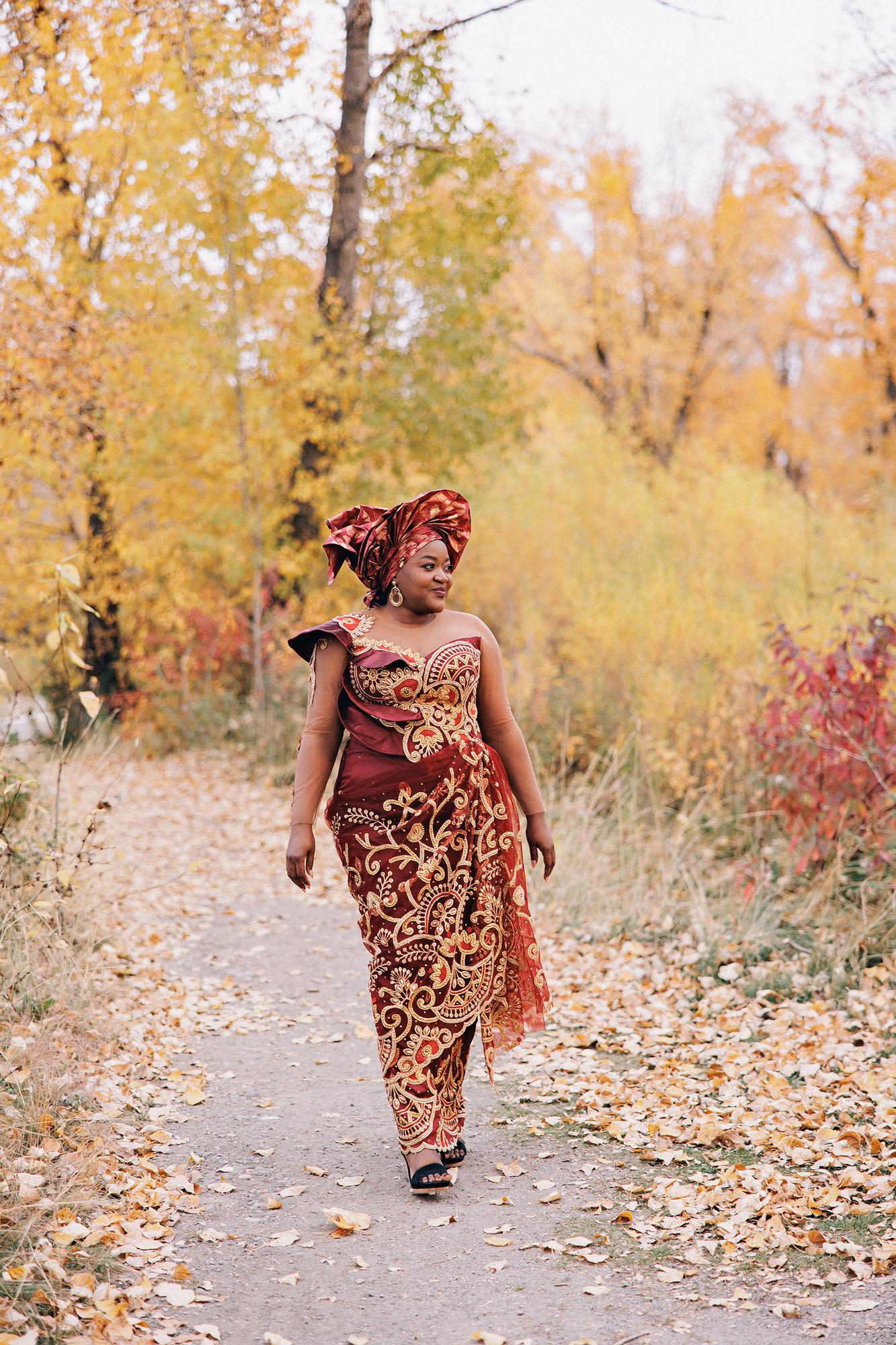 Outdoor birthday photography Calgary fall: Full-length portrait of a woman in a burgundy and gold embroidered Nigerian lace gown and gele headwrap walking confidently along a leaf-strewn autumn path, golden and red foliage surrounding her
