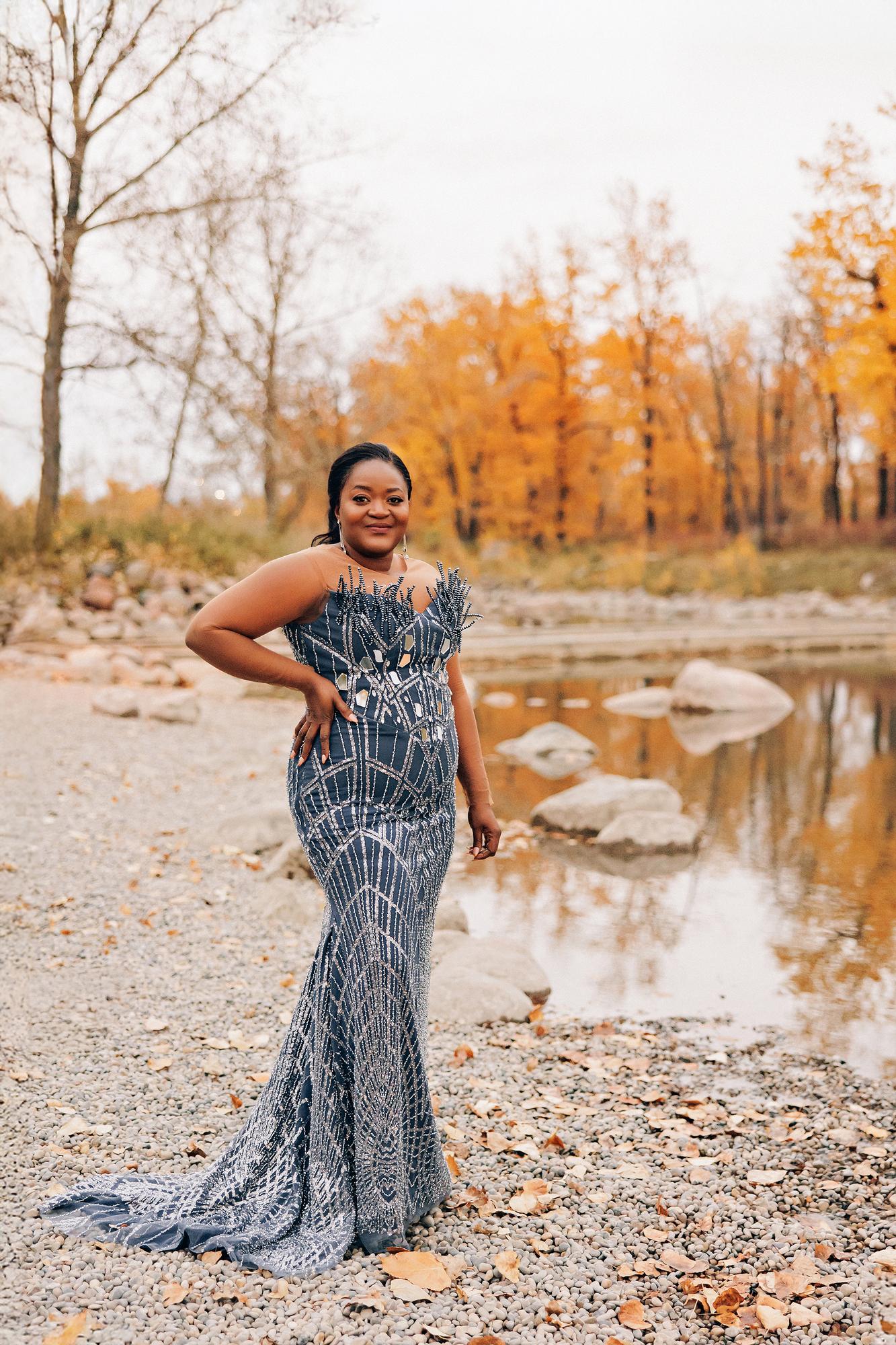 Outdoor birthday photography Calgary fall: Glamorous woman in a slate blue beaded geometric mermaid gown with mirrored embellishments and hand on hip, standing on a gravel riverbank with golden autumn trees reflected in the water behind her