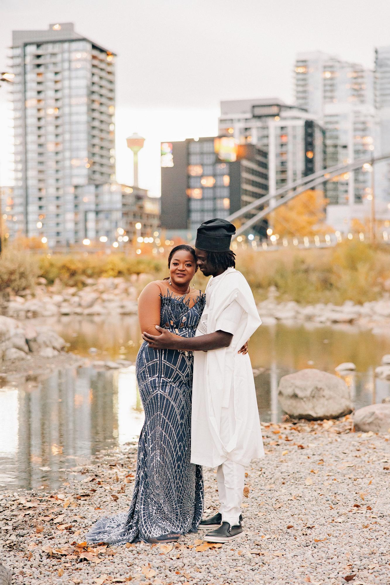 Outdoor birthday couple photography Calgary fall evening: Couple at golden hour — woman in a slate blue beaded geometric mermaid gown and man in white agbada and black fila — embracing tenderly on a gravel riverbank with the Calgary Tower and downtown skyline glowing behind them