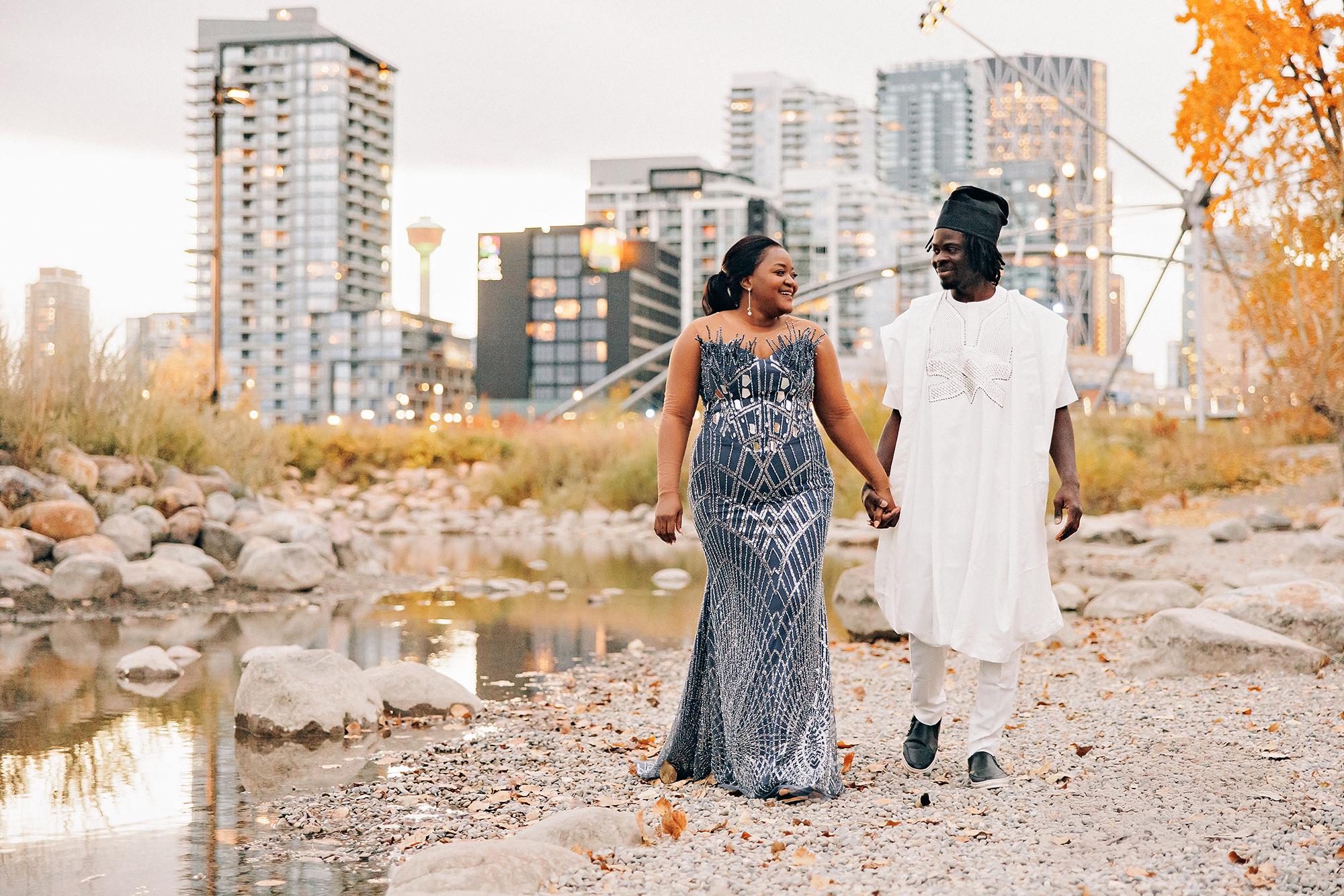 Outdoor birthday couple photography Calgary fall evening: Couple in Nigerian traditional attire walking hand-in-hand along a Bow River gravel bank at dusk — woman in a slate blue beaded gown, man in white agbada — Calgary skyline and warm city lights stretching behind them