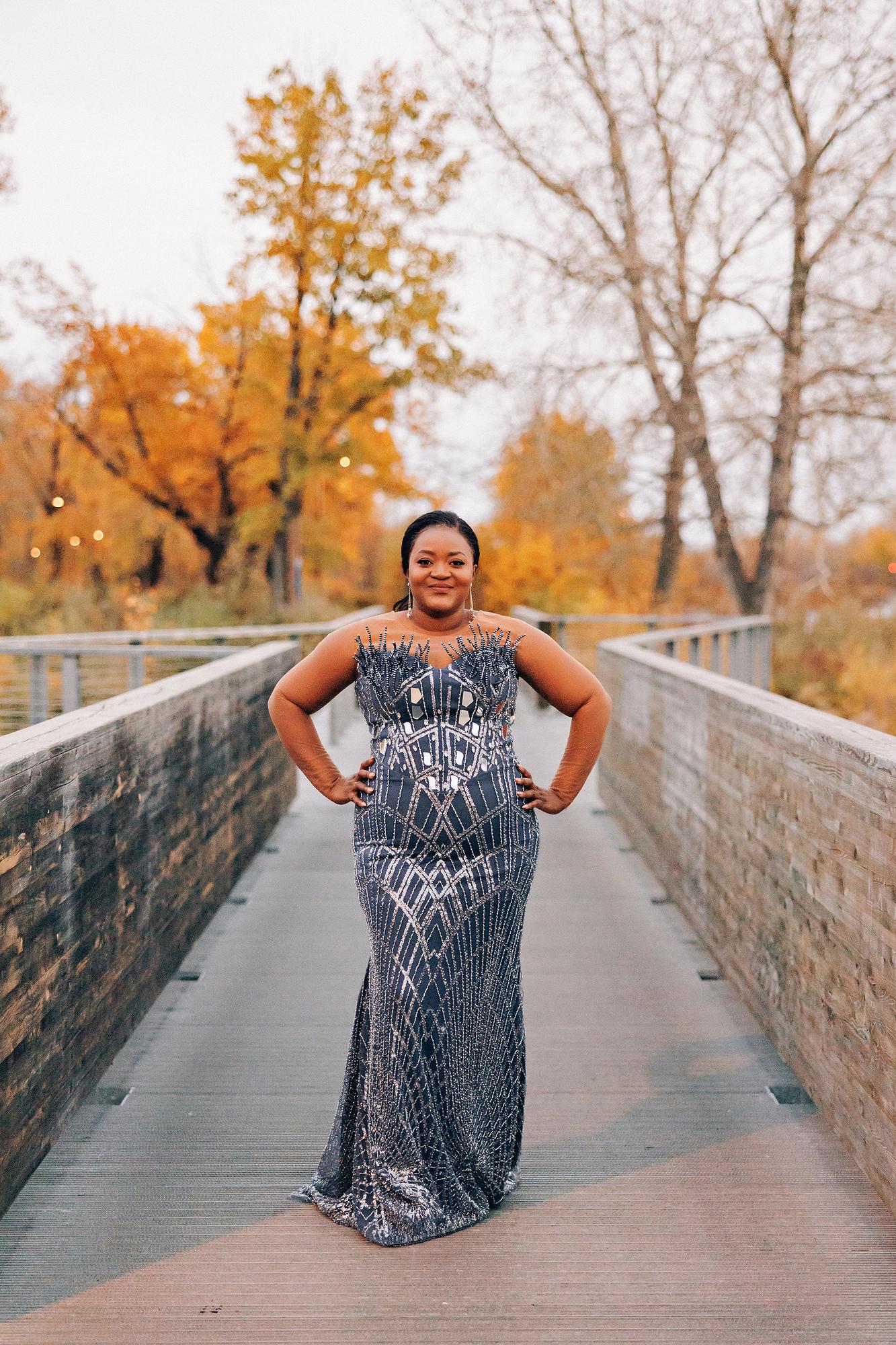 Outdoor birthday photography Calgary fall evening: Radiant woman in a slate blue beaded geometric mermaid gown with hands on hips, standing confidently on a narrow wooden bridge with golden autumn trees and soft evening light behind her