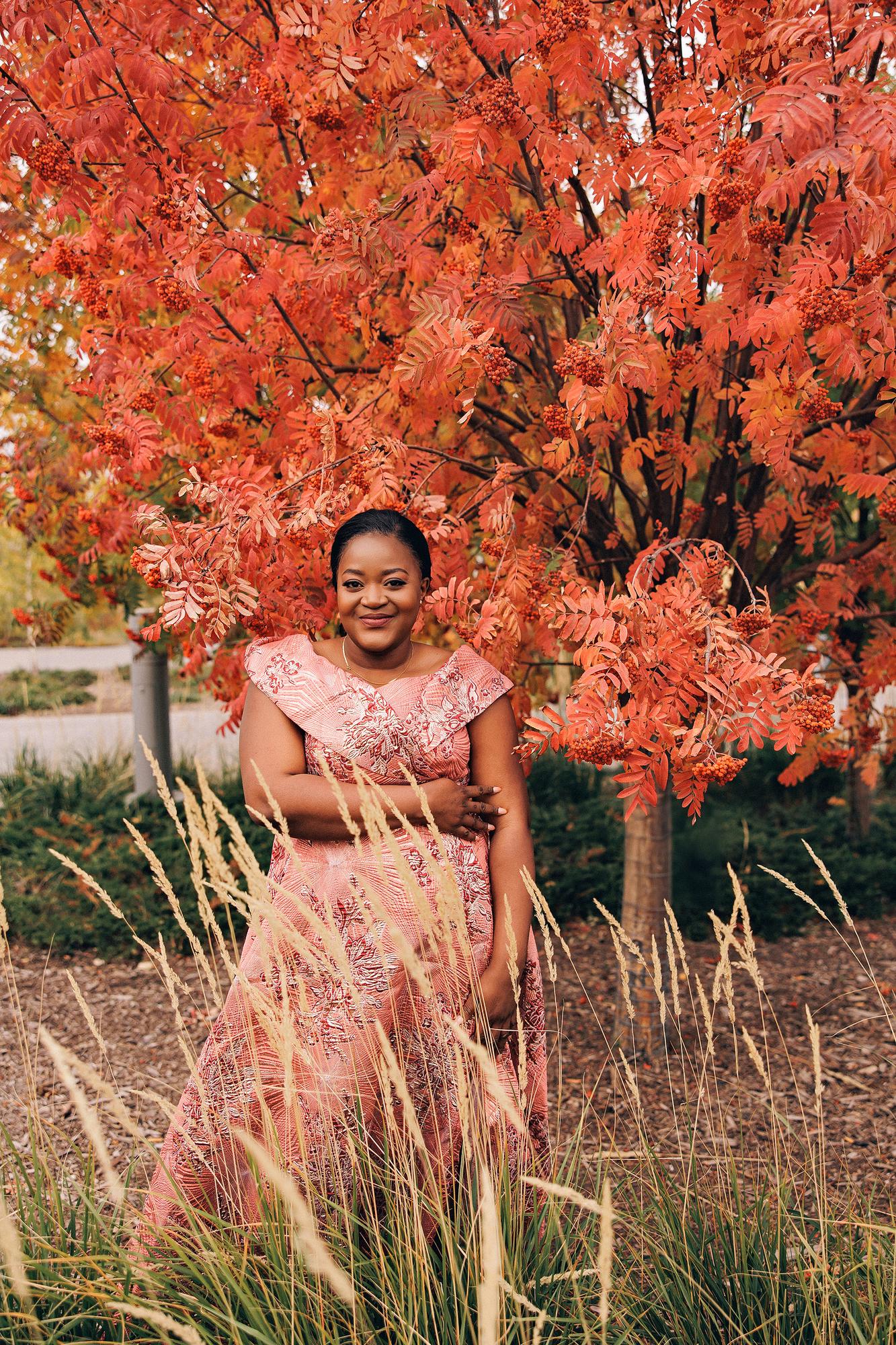 Outdoor birthday photography Calgary fall: Smiling woman in a pink-coral brocade jacquard off-shoulder gown with arms crossed, standing among golden ornamental grasses beneath a vibrant red mountain ash tree in full berry