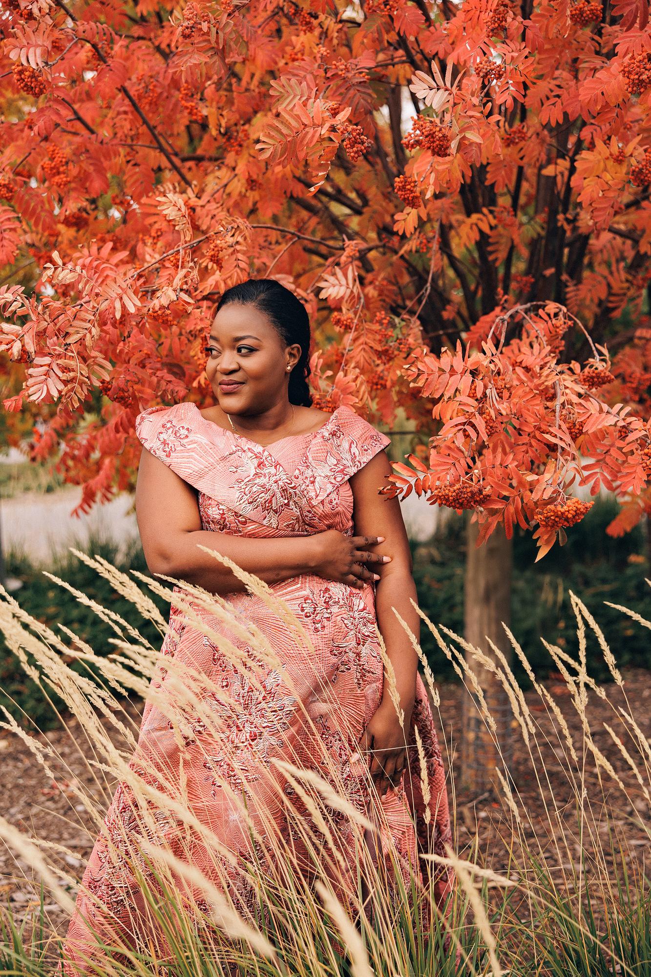 Outdoor birthday photography Calgary fall: Relaxed woman in a pink-coral brocade jacquard off-shoulder gown gazing softly to the side, framed by red-orange mountain ash foliage and dried ornamental grasses