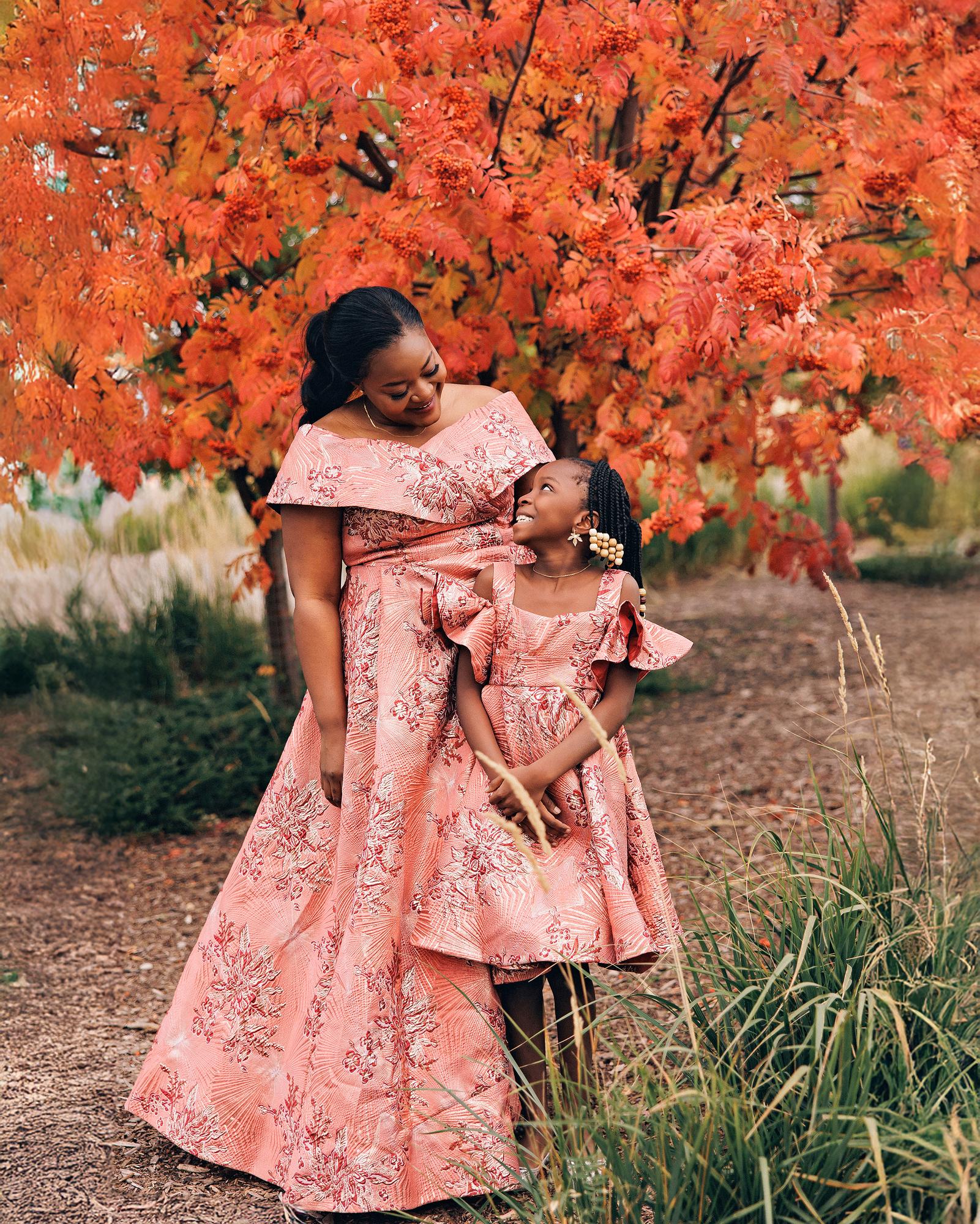 Outdoor birthday photography Calgary fall: Mother and young daughter in matching pink-coral brocade jacquard gowns — daughter in a ruffle-shoulder dress with box bow and beaded earrings — smiling warmly at each other beneath a vibrant red mountain ash tree