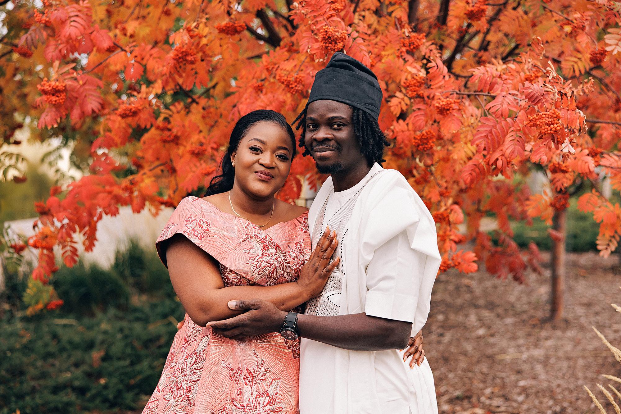 Outdoor birthday couple photography Calgary fall: Couple in coordinated Nigerian traditional attire — woman in pink-coral brocade off-shoulder gown and man in a white agbada with black fila cap — embracing and smiling at camera beneath a brilliant red mountain ash tree