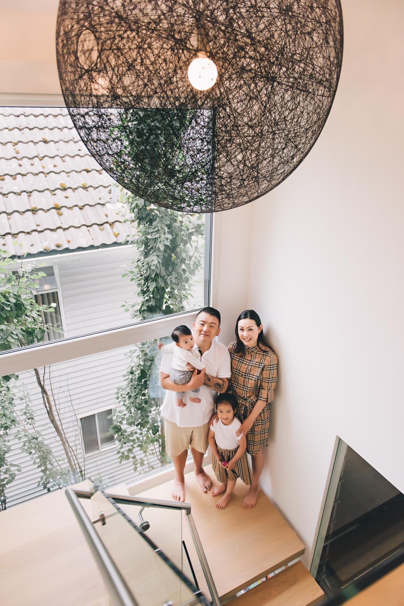 In-home lifestyle family photography: Overhead portrait of a family of four on a bright staircase landing — father in a white shirt holding a baby boy, mother in a Burberry plaid dress with young daughter in a white top — a large wire globe pendant light above them, floor-to-ceiling window with lush greenery behind them