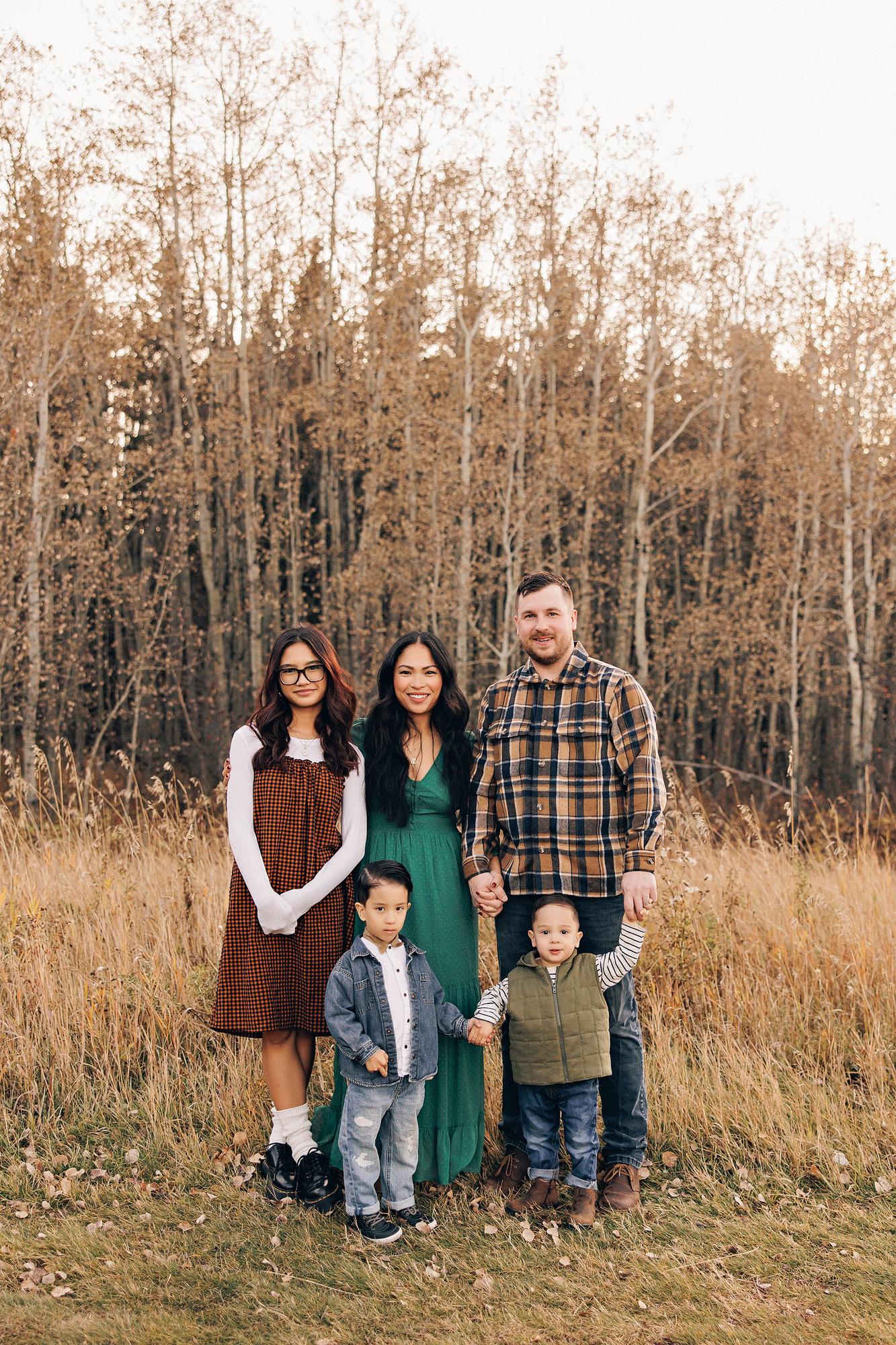 Outdoor family photography Calgary fall: Family of five — mother in an emerald green maxi dress, father in a brown plaid flannel shirt, teenage daughter in an orange gingham dress with bold black-frame glasses, and two young boys in denim and olive — standing together in a golden meadow beside a birch forest