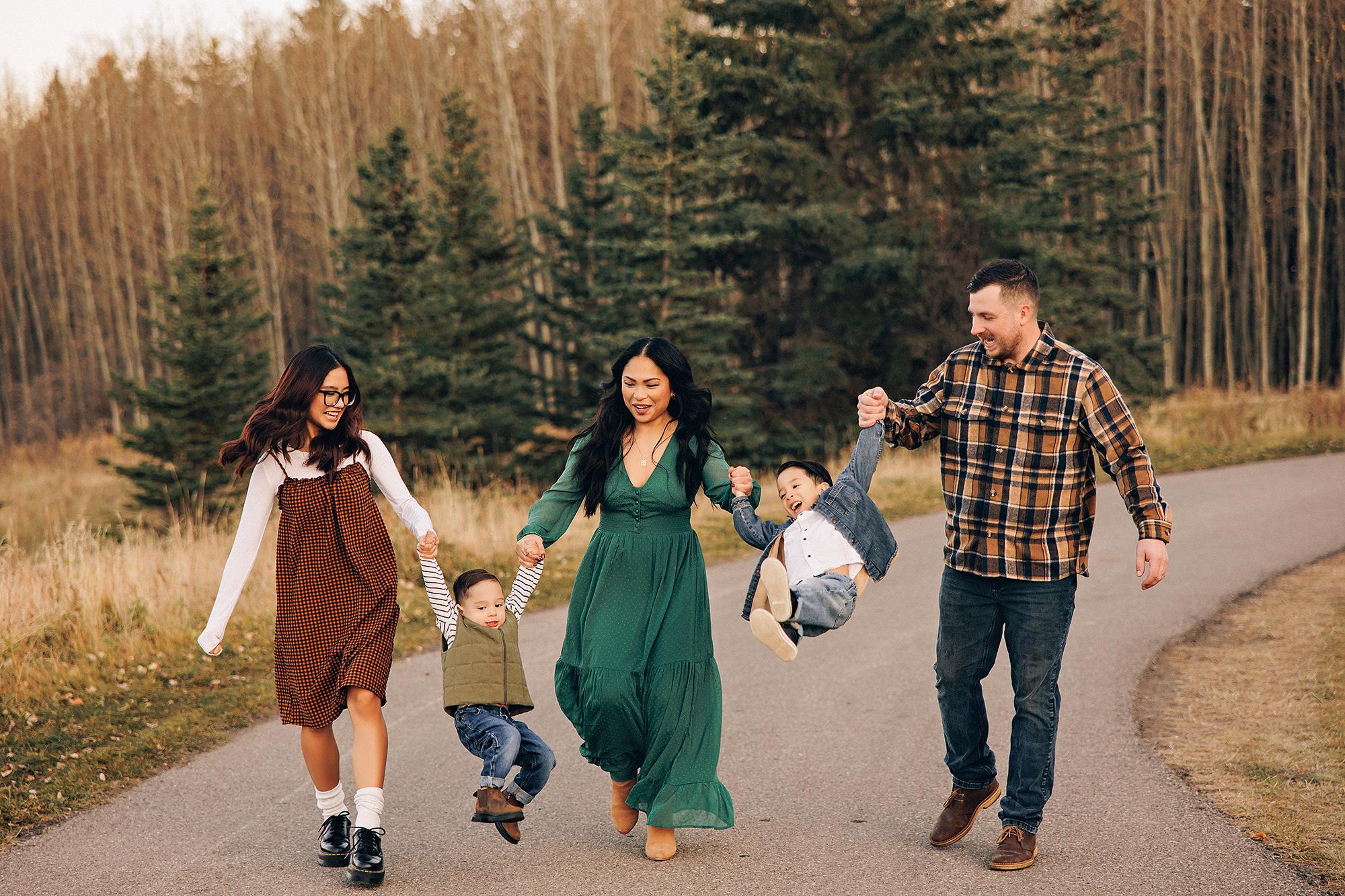 Outdoor family photography Calgary fall: Father in brown plaid flannel crouching with arms around two grinning young sons in a golden meadow — toddler in olive vest and striped top, older boy in denim jacket — warm autumn birch forest behind them