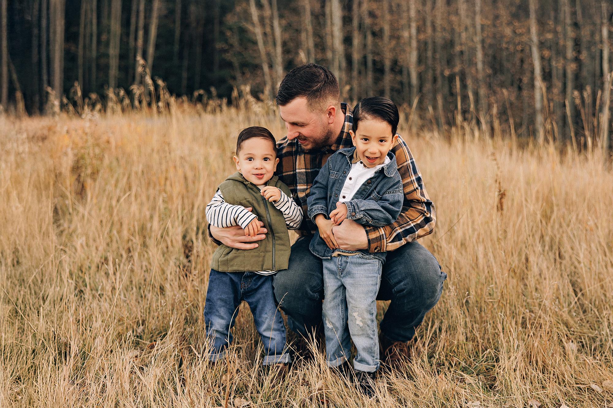 Outdoor family photography Calgary fall: Joyful family of five walking along a paved park path — parents and teenage daughter in orange gingham swinging two laughing young boys by the arms, tall evergreen and bare aspen trees lining the path behind them