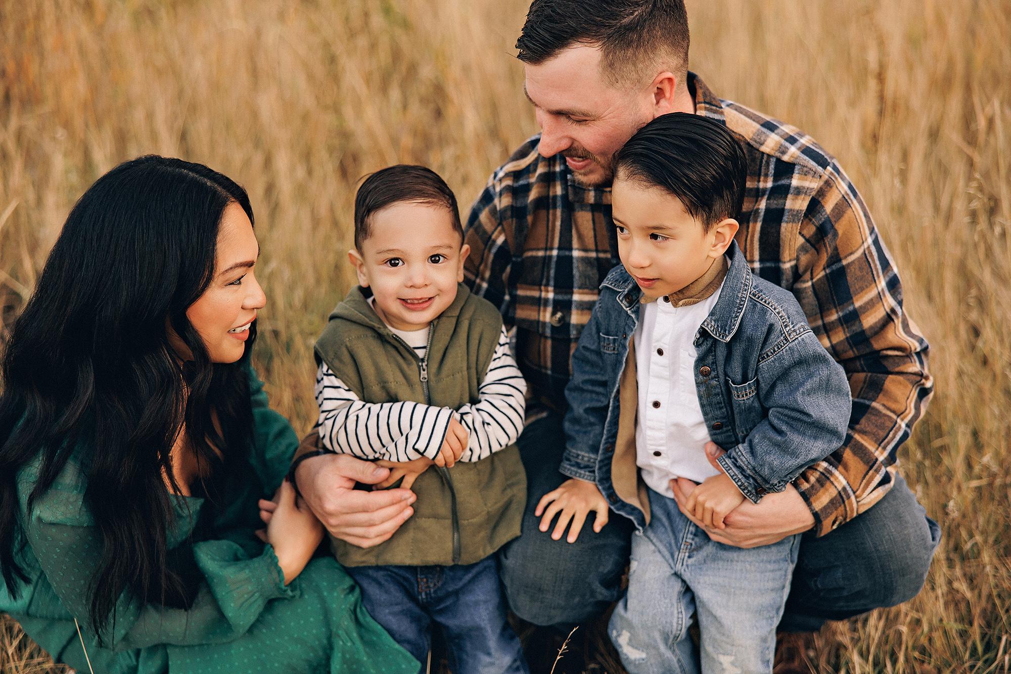Outdoor family photography Calgary fall: Intimate close-up of parents crouched in golden field grass with their two young sons — mother in emerald green dress smiling at toddler, father in plaid flannel with arm around older boy in denim jacket