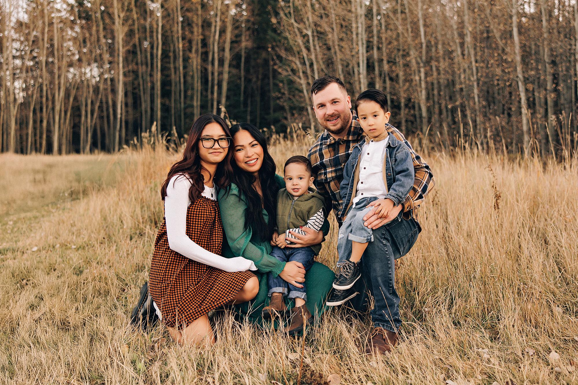 Outdoor family photography Calgary fall: Family of five seated together in a golden meadow at dusk — mother in emerald green maxi dress with toddler in her lap, teenage daughter in orange gingham beside her, father in plaid flannel with older son — aspen forest backdrop