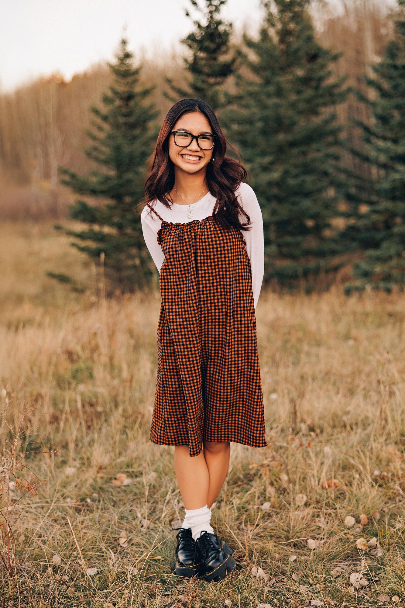 Outdoor portrait photography Calgary fall: Radiant teenage girl with dark wavy auburn-highlighted hair and bold black-frame glasses in an orange gingham ruffle sundress over a white long-sleeve top, smiling brightly in front of tall evergreen trees
